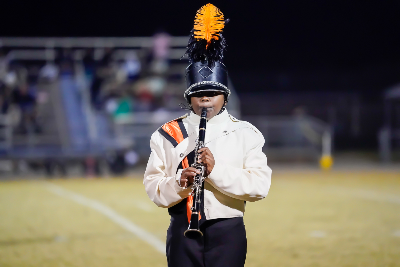 A marching band member in a white and black uniform with an orange and black plume on their hat plays a clarinet on a field at night. The background is blurred, showing some bleachers and people.