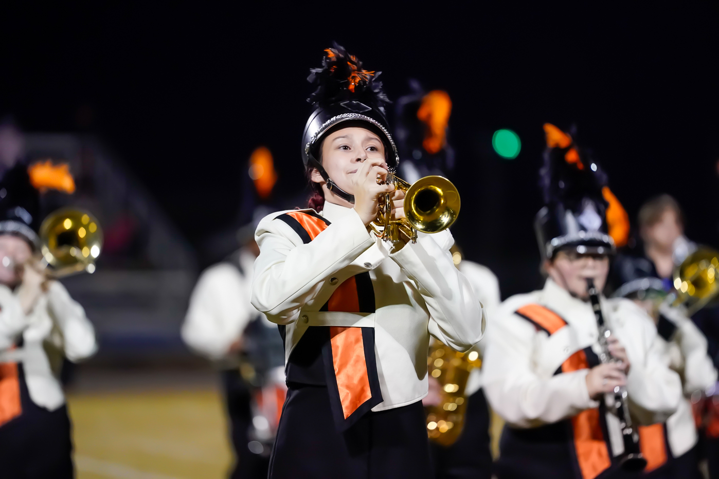 A marching band member in a black and white uniform with orange accents plays a trumpet at night, surrounded by other band members holding instruments.