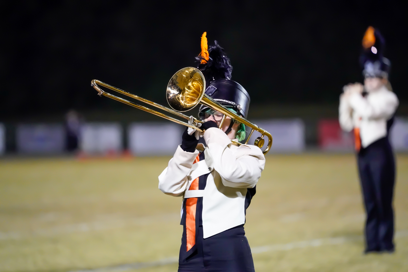 A marching band member in uniform plays a trombone on a field at night, with another band member blurred in the background.