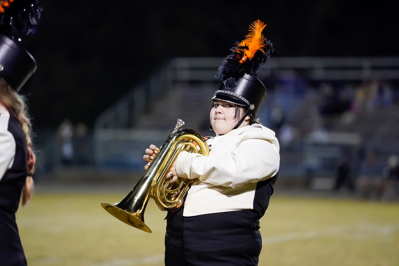A marching band member in a black and white uniform and a tall hat with orange and black plumes plays a brass instrument on a field, with blurred bleachers in the background.