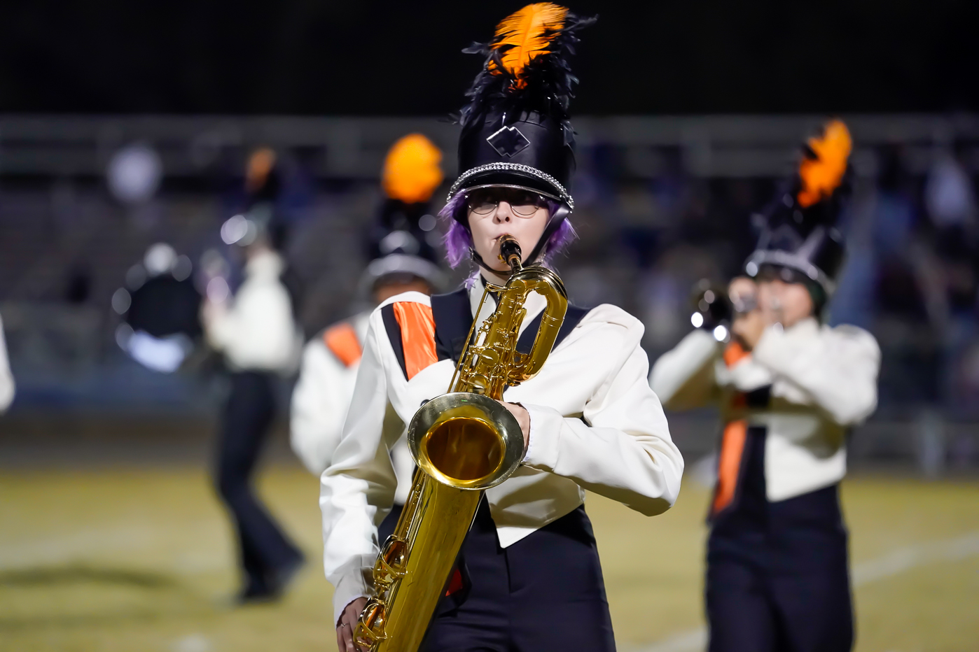 A marching band member in a white and black uniform with orange accents plays a saxophone on a field. The person wears a tall hat decorated with orange and black feathers. Other band members are visible in the background.