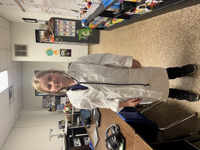 A young girl wearing a white lab coat and stethoscope stands in a classroom setting with various educational materials and toys visible in the background.