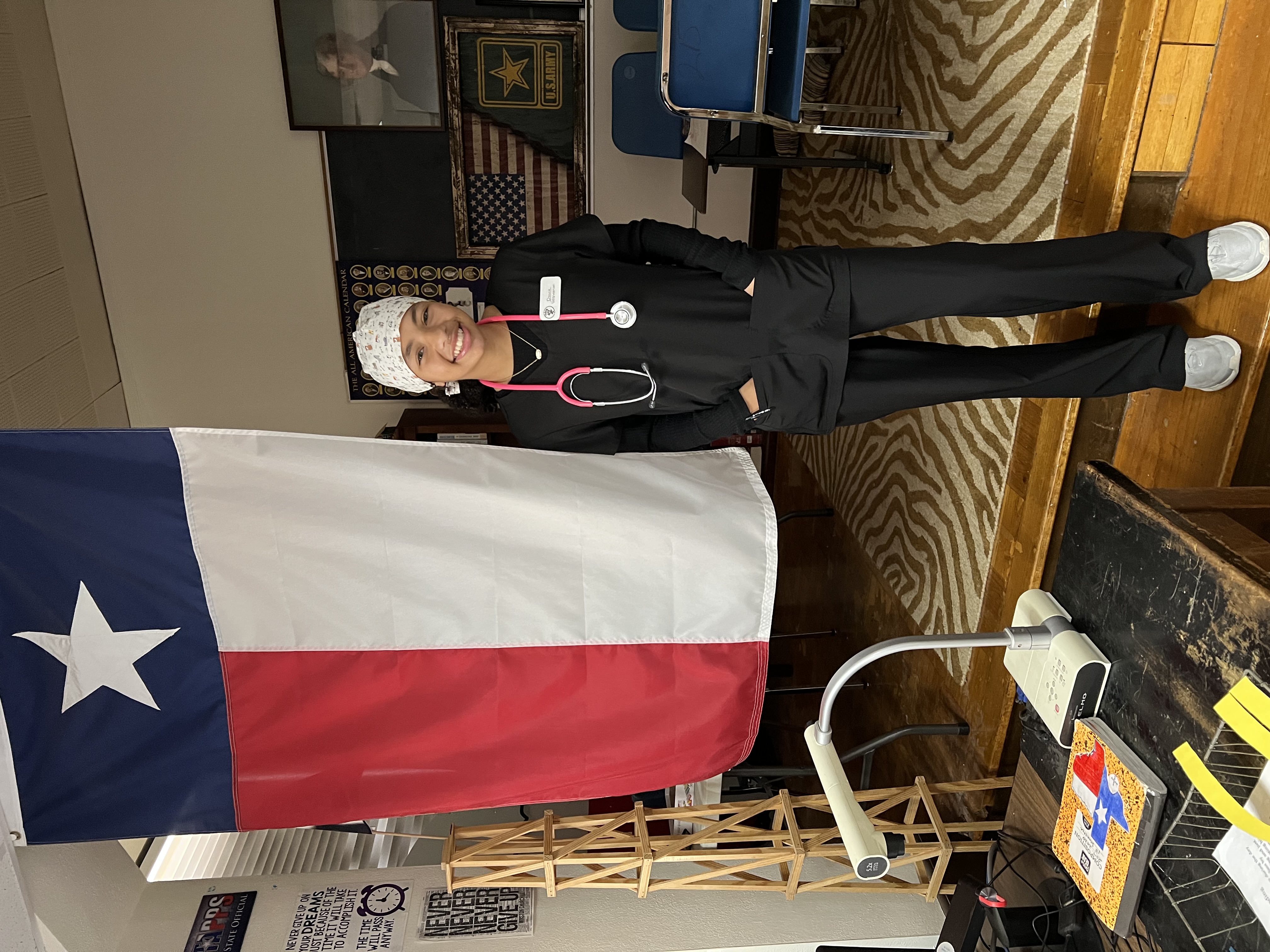 A person wearing a white medical coat and hat stands in front of a large Texas state flag in what appears to be a classroom or medical setting.
