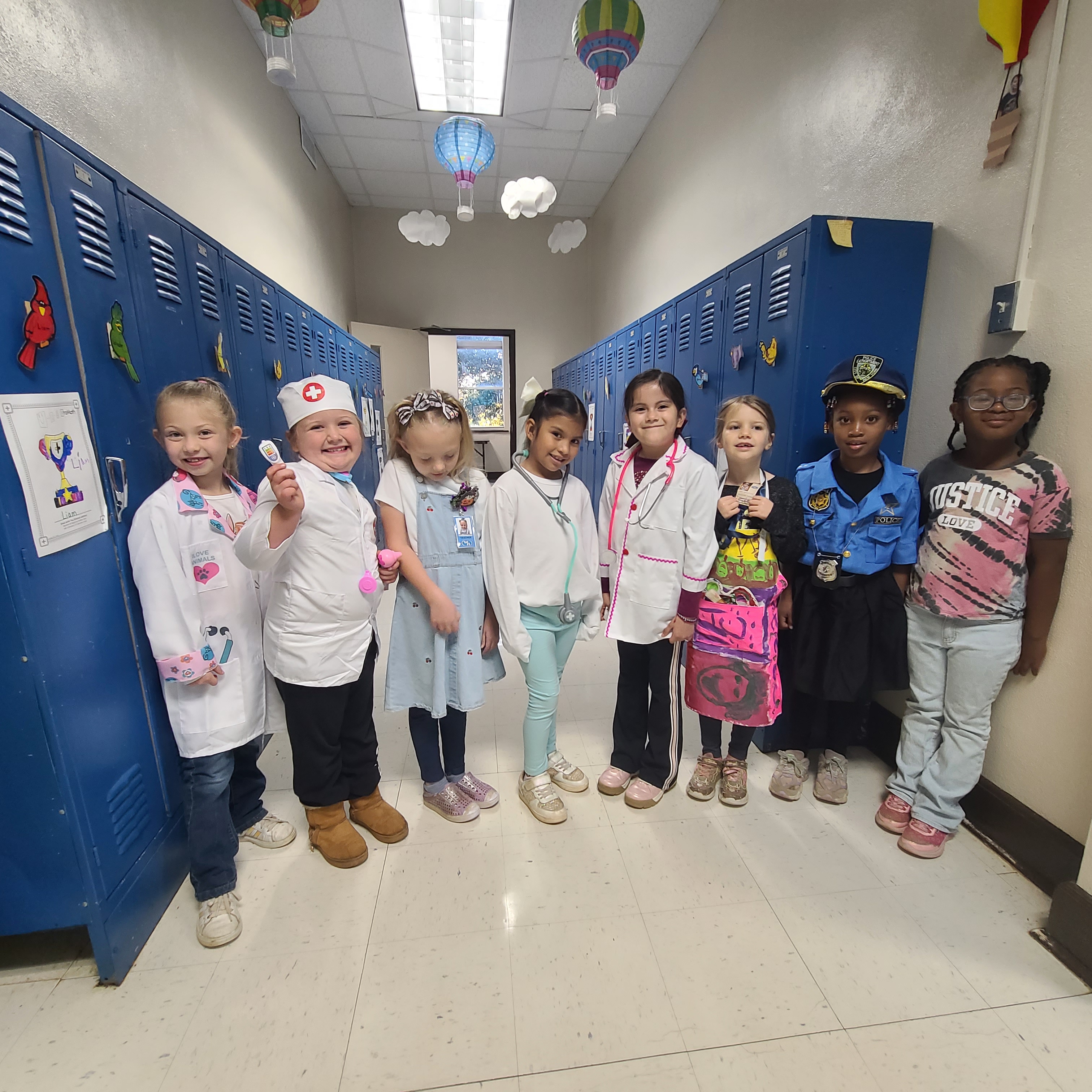 A group of young children, dressed in various costumes, stand together in a school hallway with blue lockers and colorful decorations in the background.
