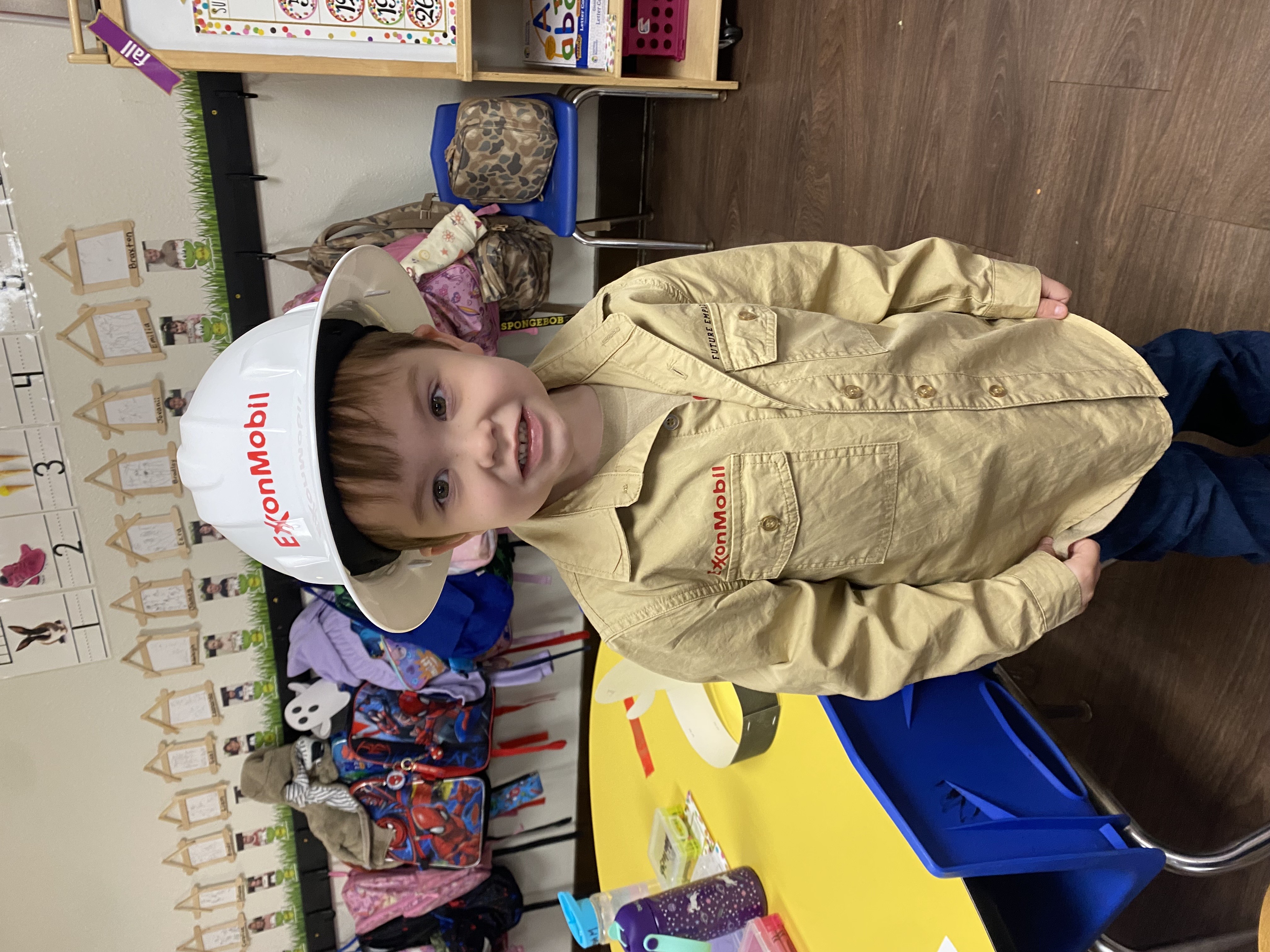 A young boy wearing an Exxon Mobil hat and a beige shirt stands in a colorful classroom setting with various toys and learning materials visible in the background.