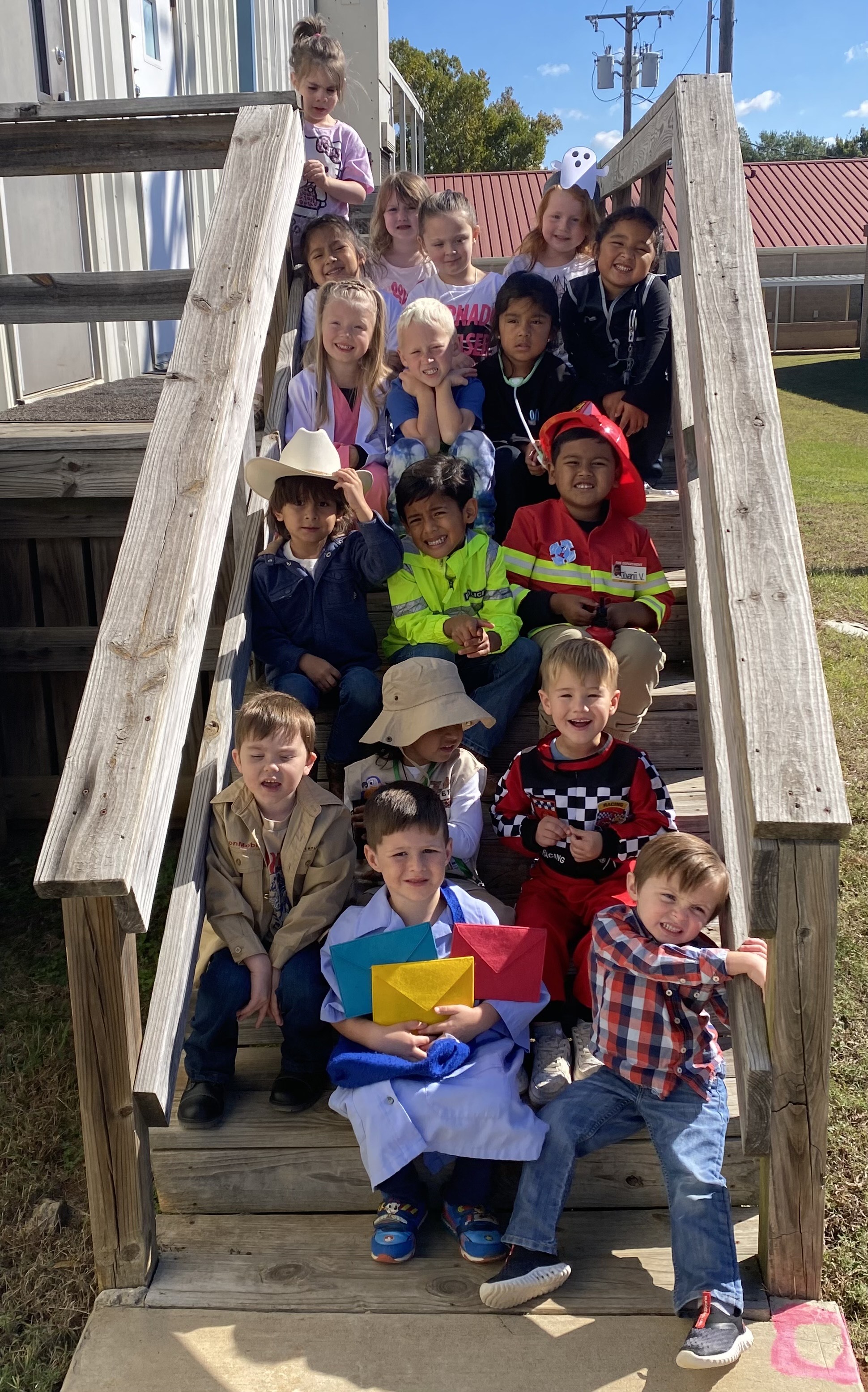 A group of young children, both boys and girls, sitting on wooden steps outdoors, surrounded by a building and a grassy area in the background.