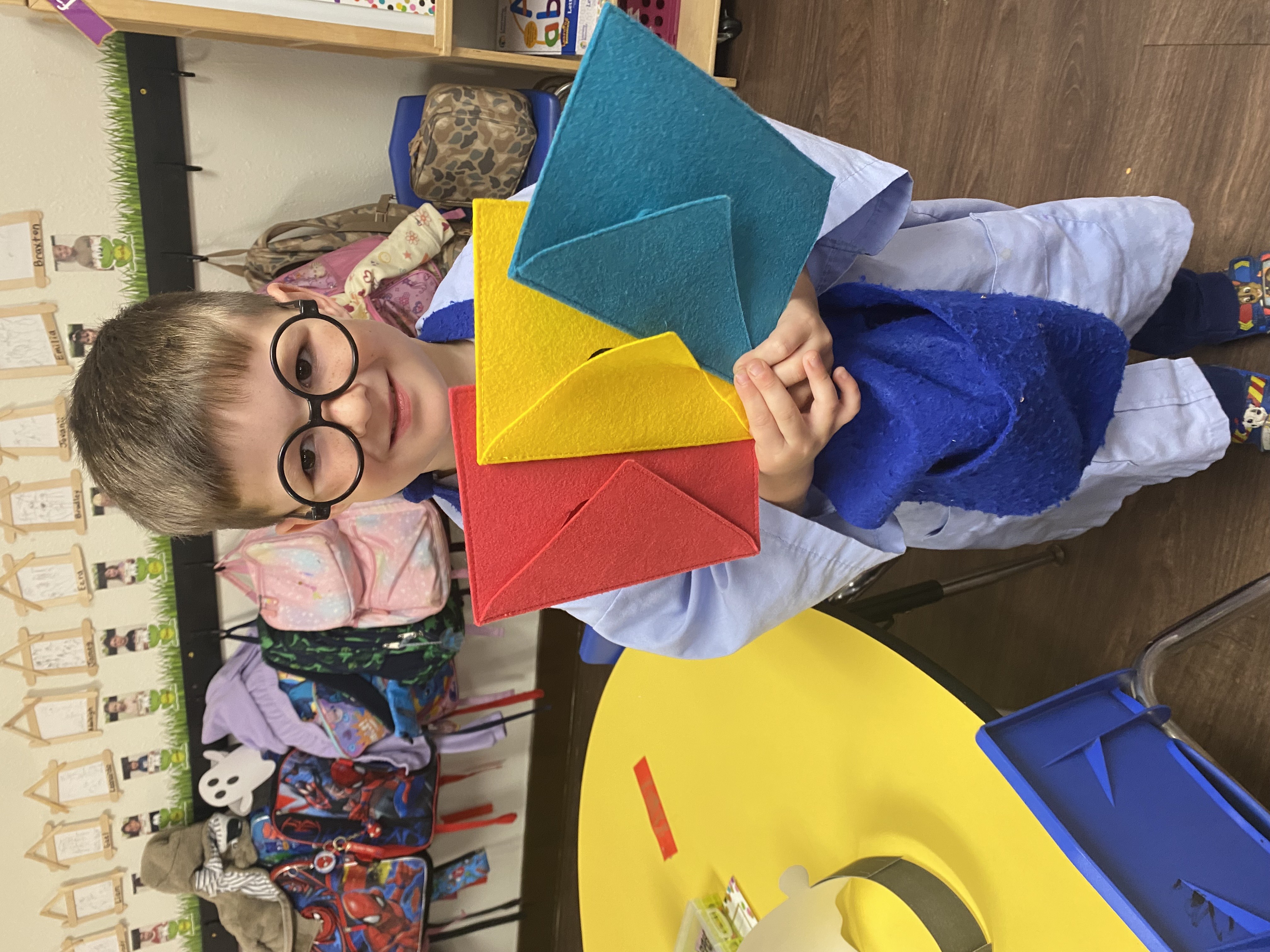 A young boy with glasses is holding colorful paper shapes in a classroom setting with backpacks and other school supplies visible in the background.