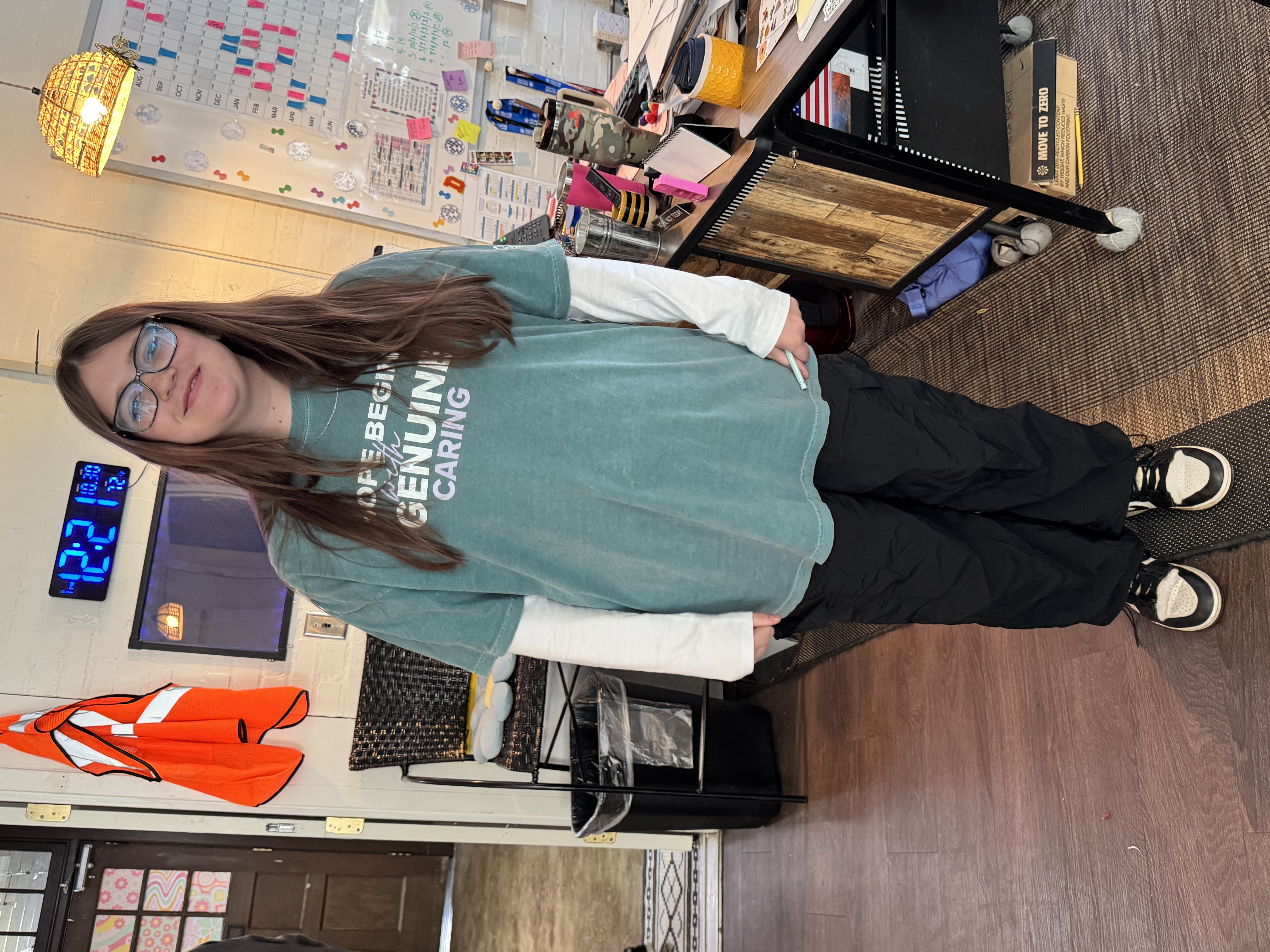 The image shows a young woman wearing a green sweatshirt with the text "Genuine Caring" on it, standing in what appears to be a classroom or office setting with various items and furniture visible in the background.