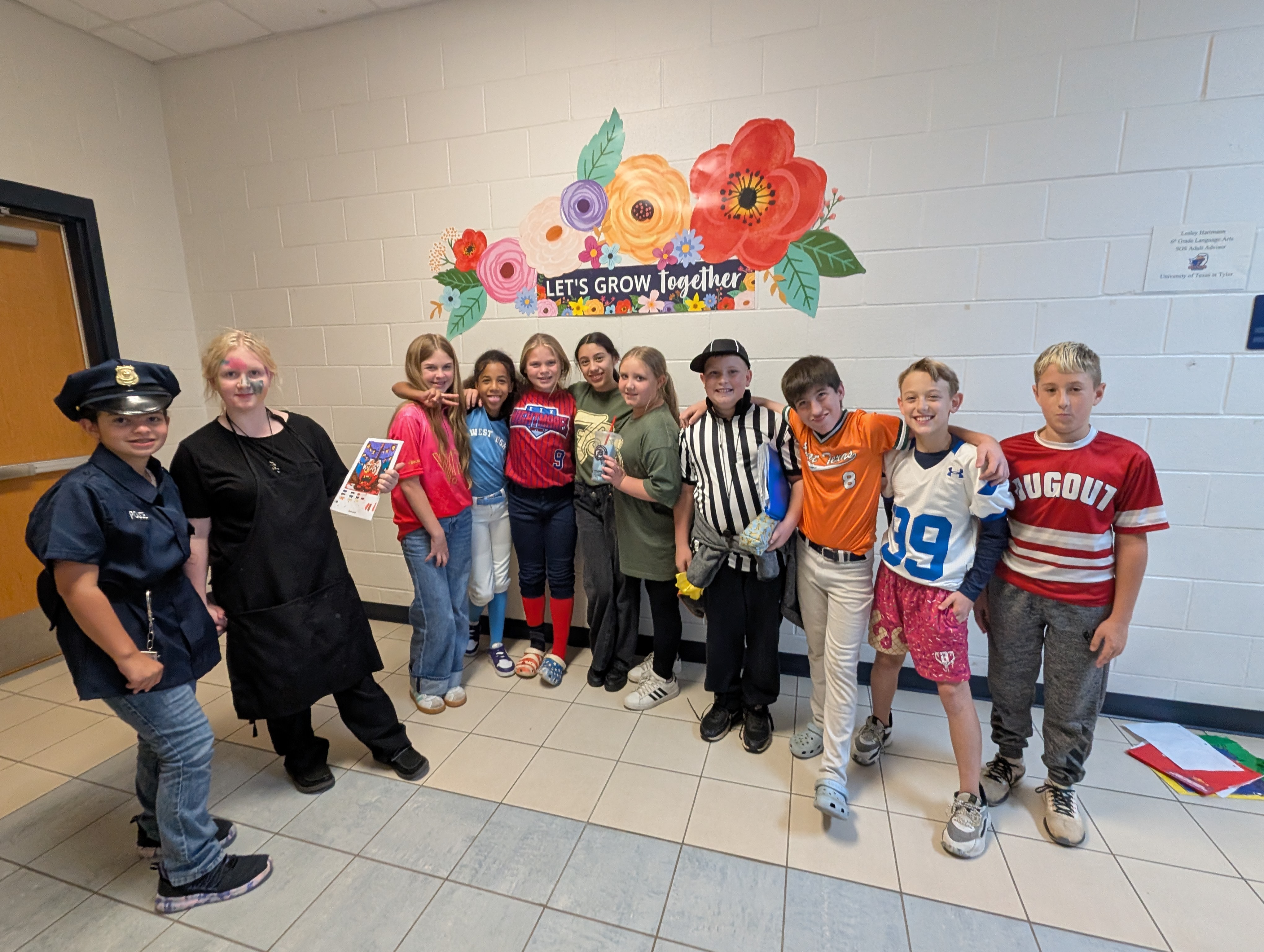 A group of children, some wearing colorful clothing, stand together in front of a wall decorated with a large, vibrant floral mural with the text "Let's grow together".