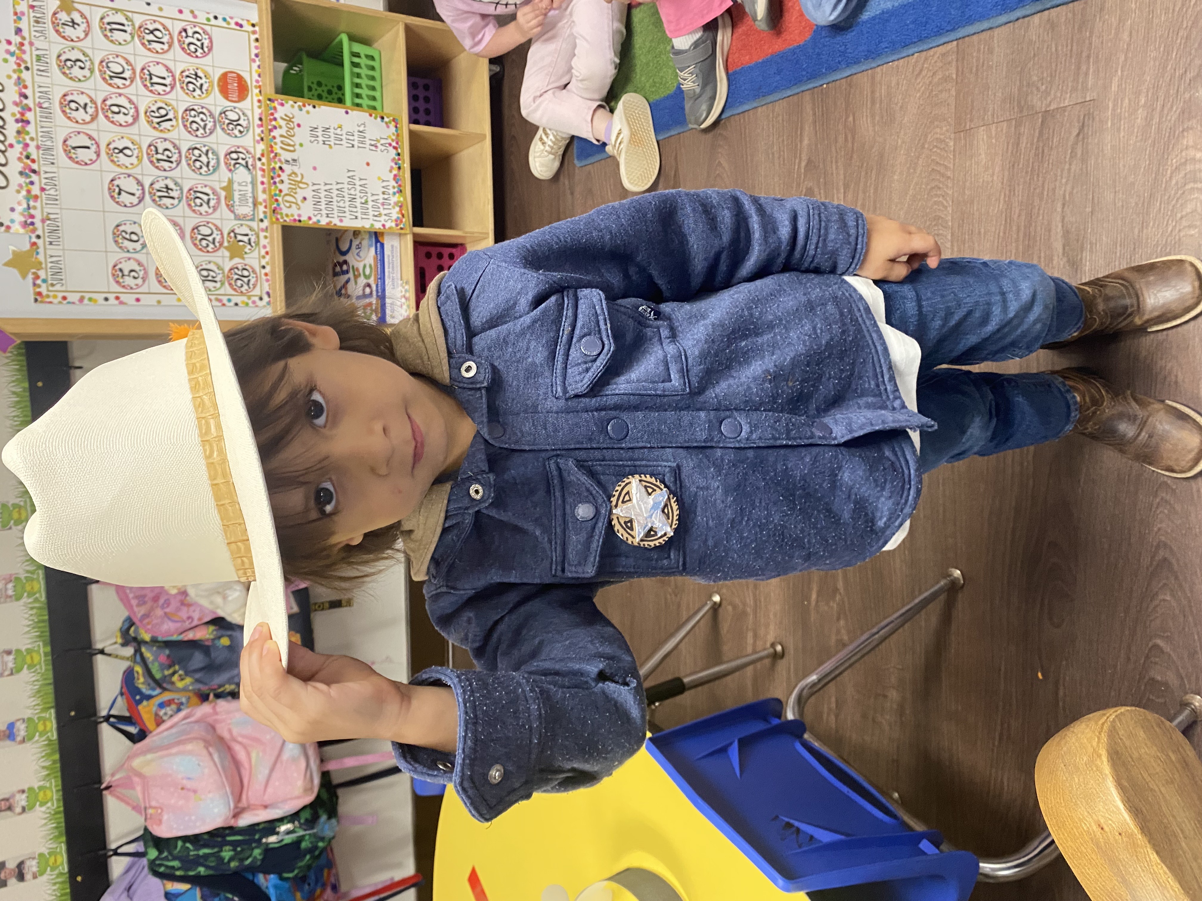 A young child wearing a denim jacket and cowboy hat stands in a classroom setting, surrounded by educational materials and furniture.