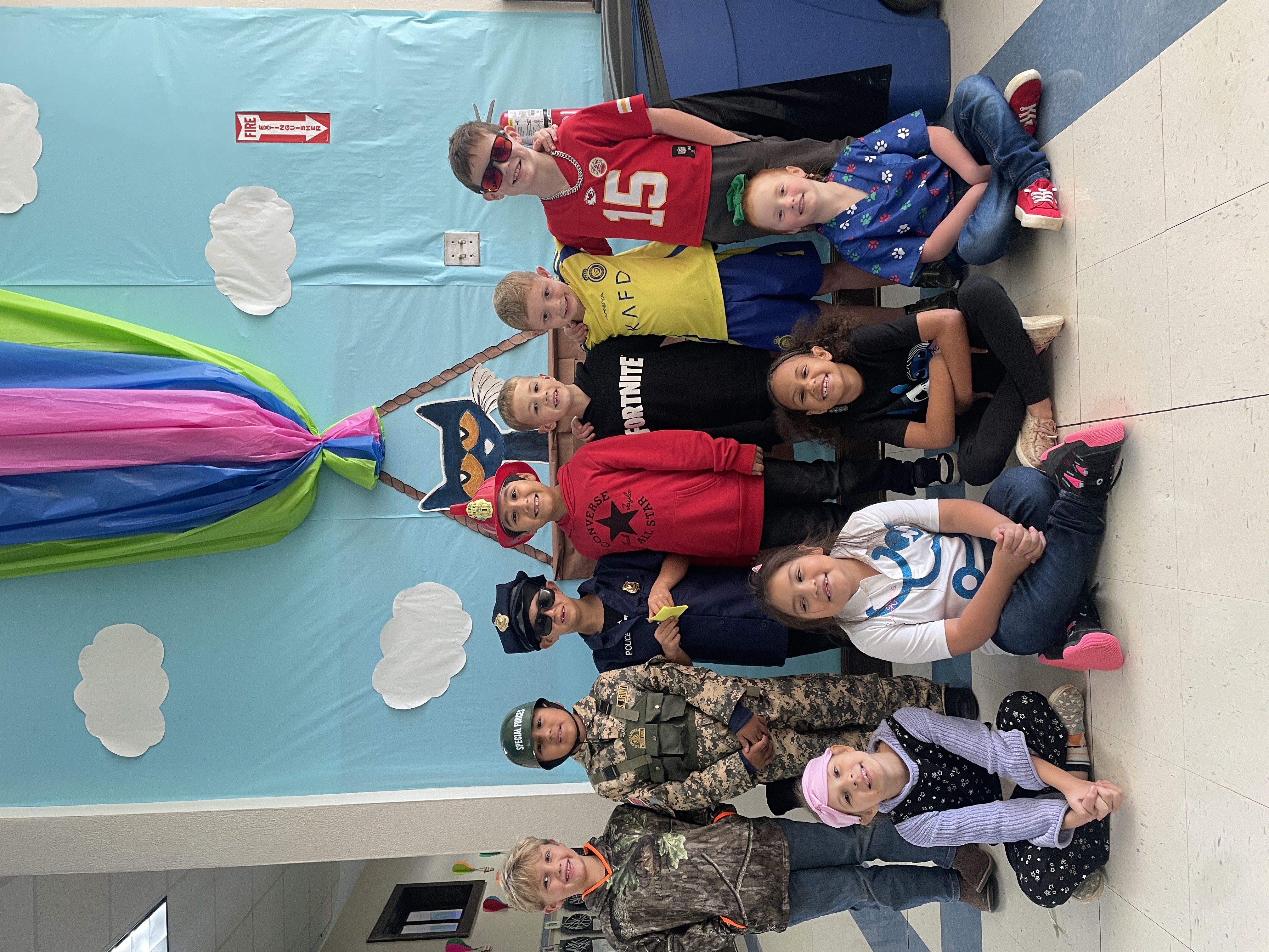 A group of children dressed in various costumes, including superheroes and military uniforms, sitting together in a classroom setting with a colorful backdrop of clouds and a parachute.