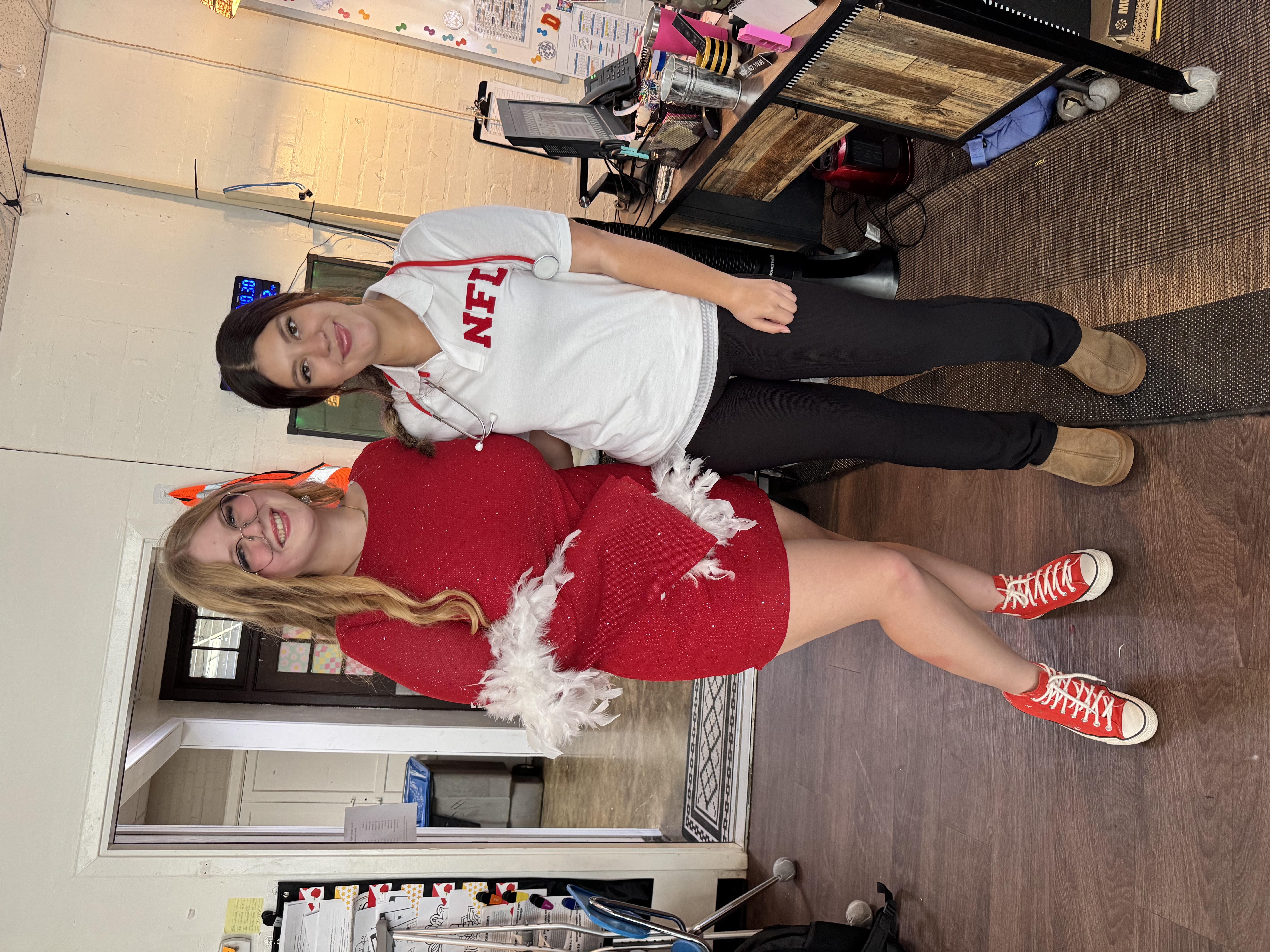 Two young women, one wearing a red outfit and the other wearing a white NFL shirt, are standing together in what appears to be a room or office setting.