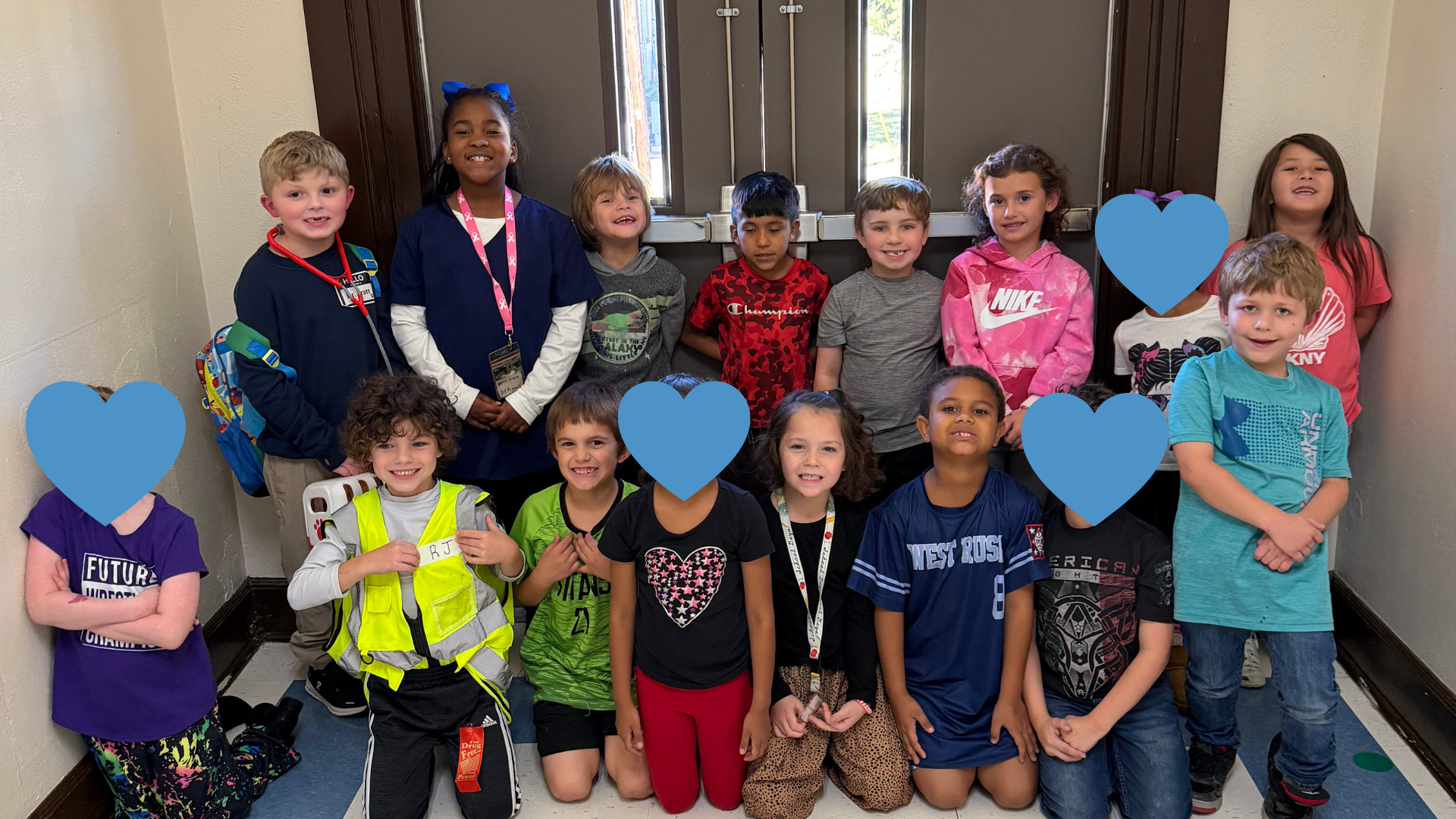 A group of young children, both boys and girls, standing together in a hallway with blue heart-shaped decorations on the walls.