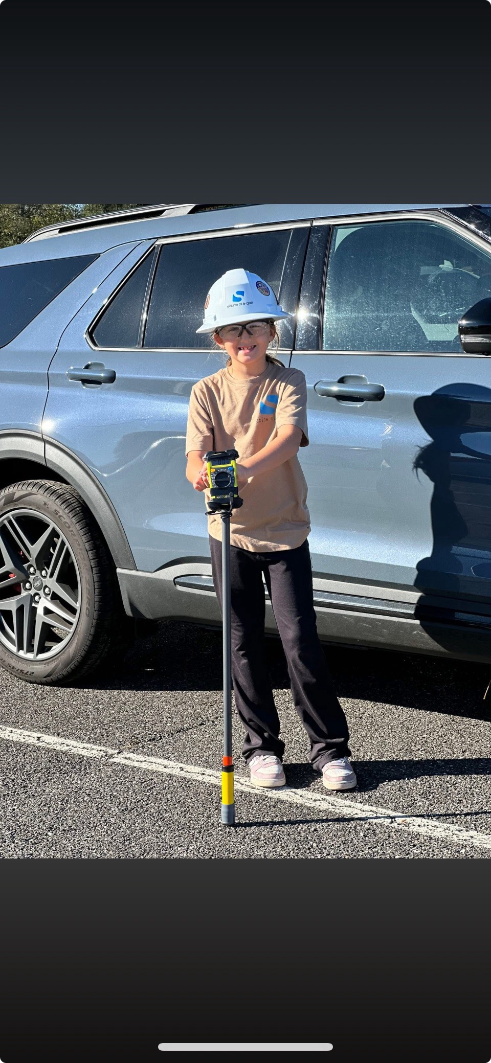 A person wearing a baseball cap and casual clothing stands in front of a large silver SUV on a paved road, with a grassy area visible in the background.