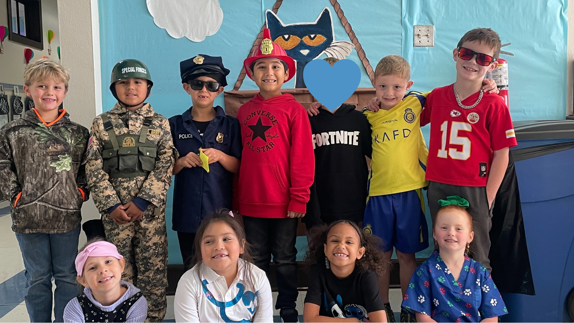 A group of children dressed in various costumes, including military uniforms, superheroes, and sports jerseys, posing together in front of a colorful backdrop with clouds and other decorations.