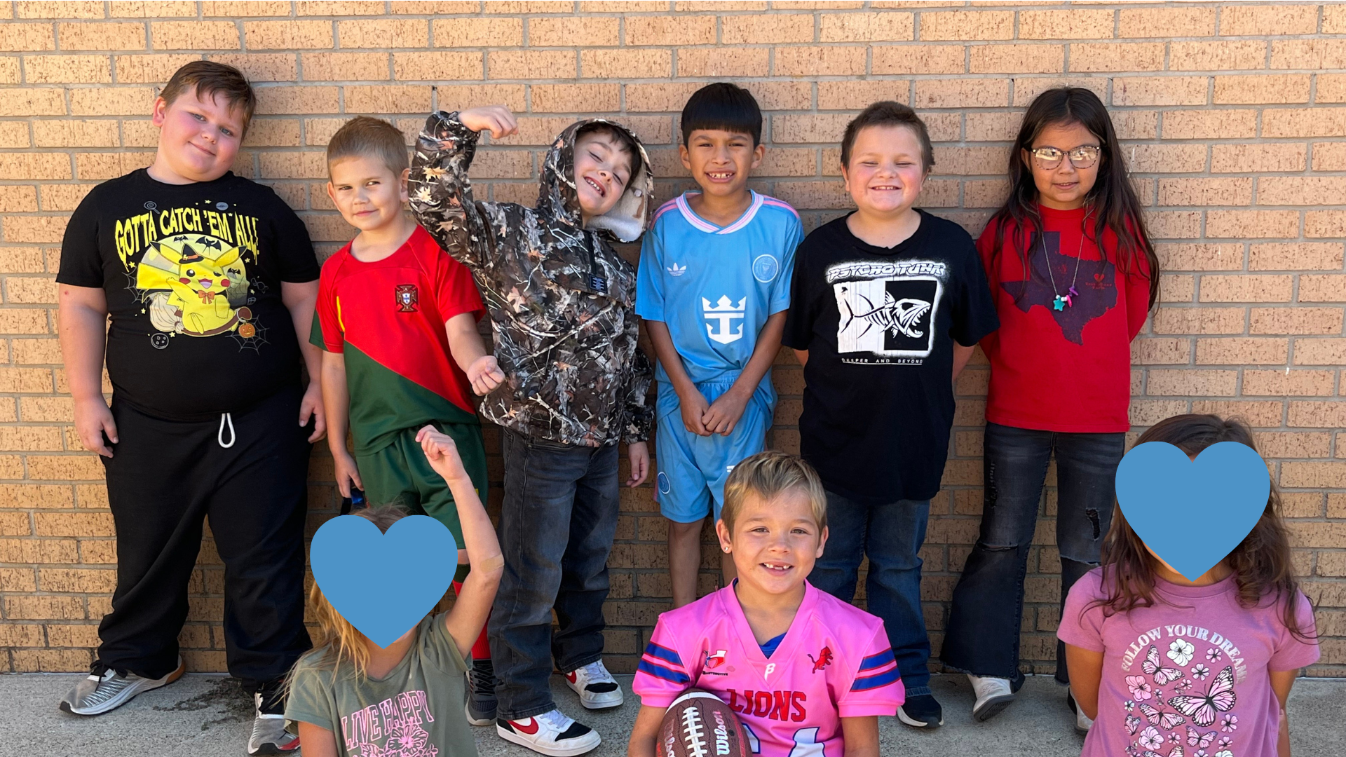 A group of young people, some wearing colorful t-shirts, stand in front of a brick wall, with blue heart-shaped cutouts in the foreground.