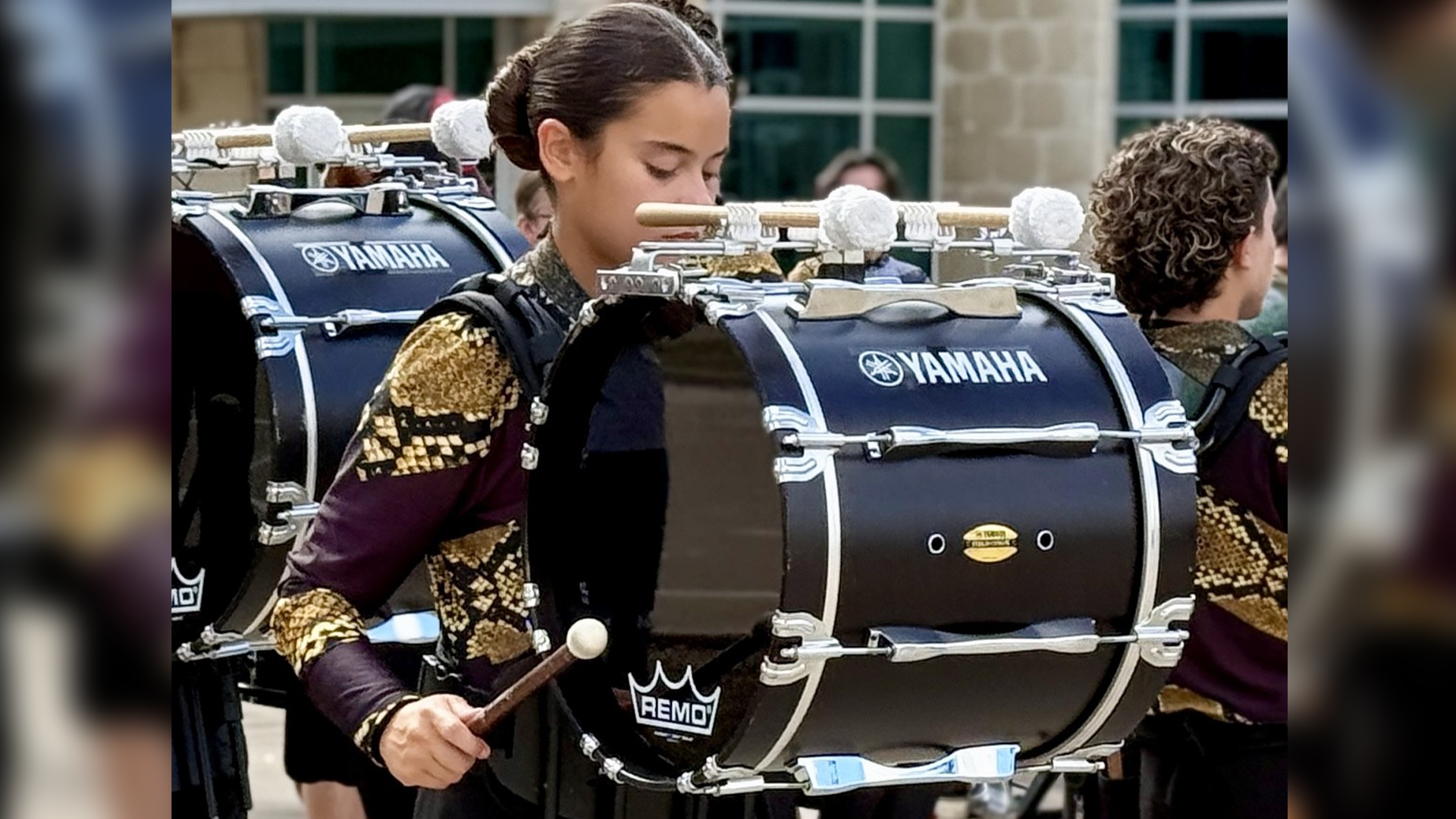 Two individuals wearing colorful, patterned clothing are playing Yamaha drum sets in what appears to be a performance or rehearsal setting.