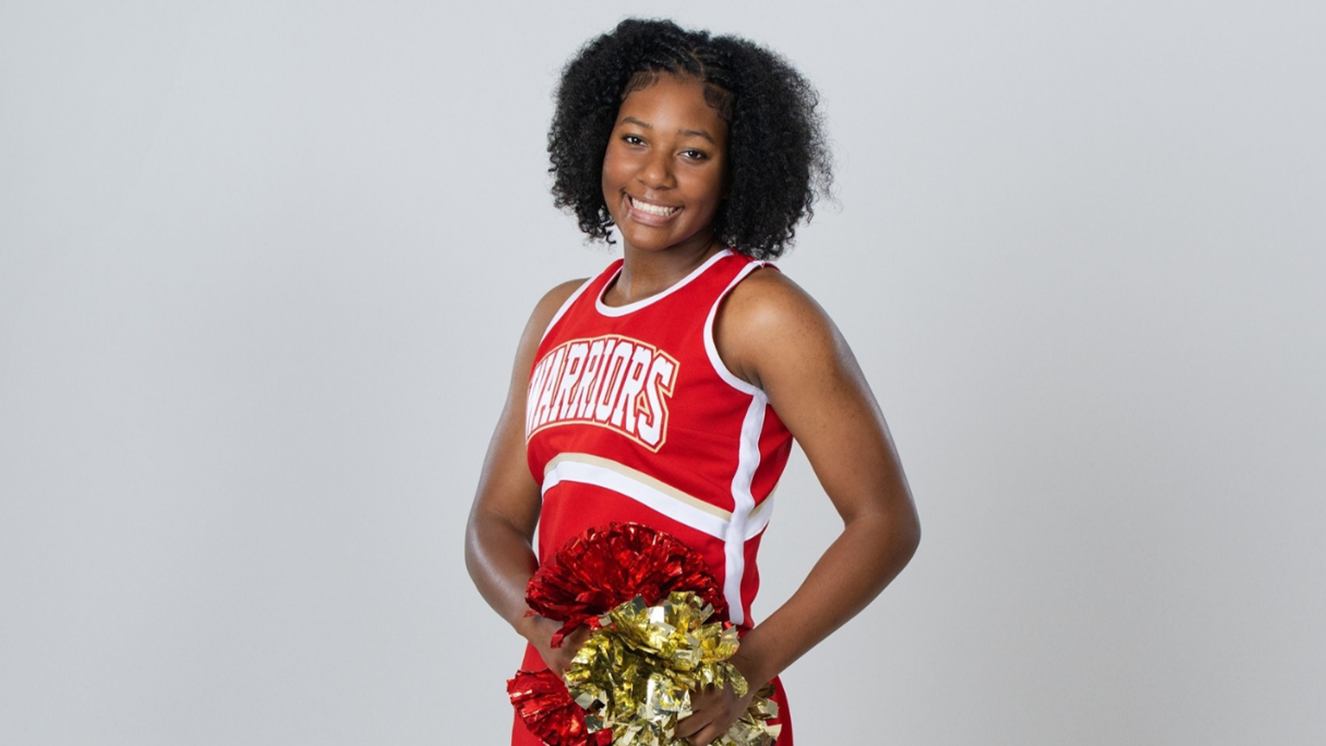 A young woman with curly dark hair wearing a red and white basketball jersey stands against a plain white background, smiling confidently.