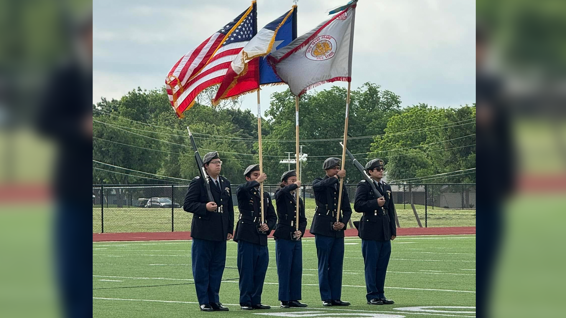A group of people in formal uniforms standing in front of American and state flags on a sports field.