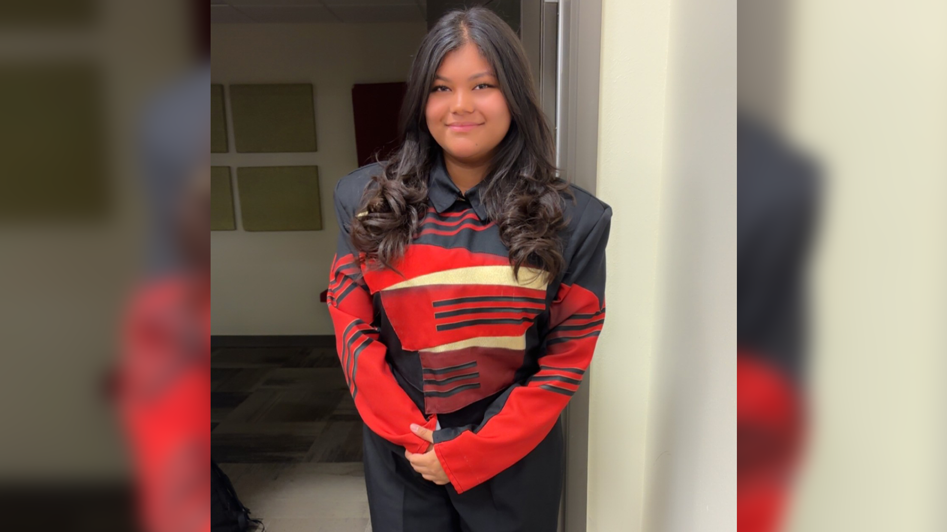 The image shows a young woman with long, dark curly hair wearing a red and black striped sweater, standing in a doorway with a neutral expression on her face.
