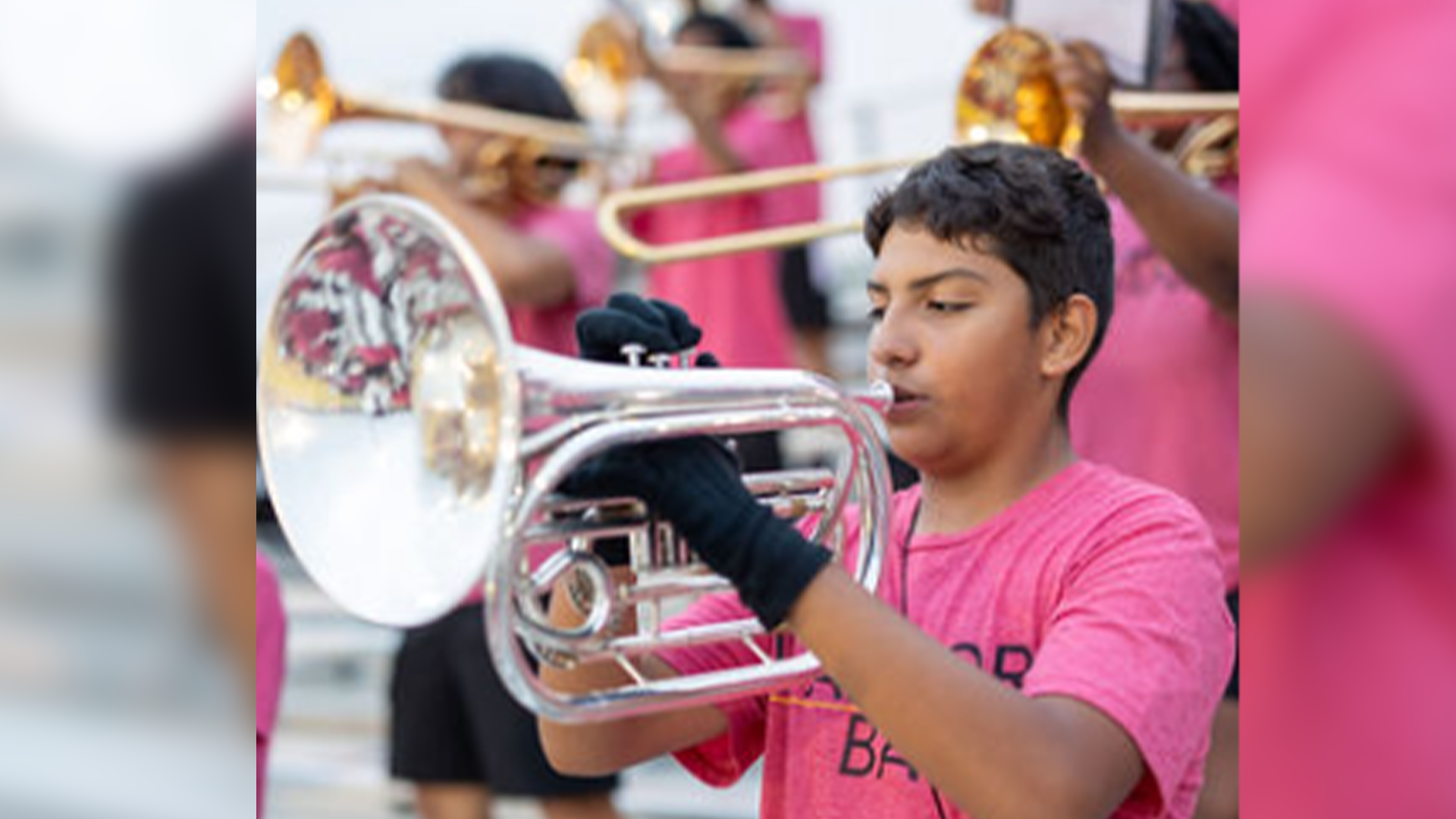 A young person in a pink shirt is playing a brass instrument, surrounded by other musicians in a performance setting.