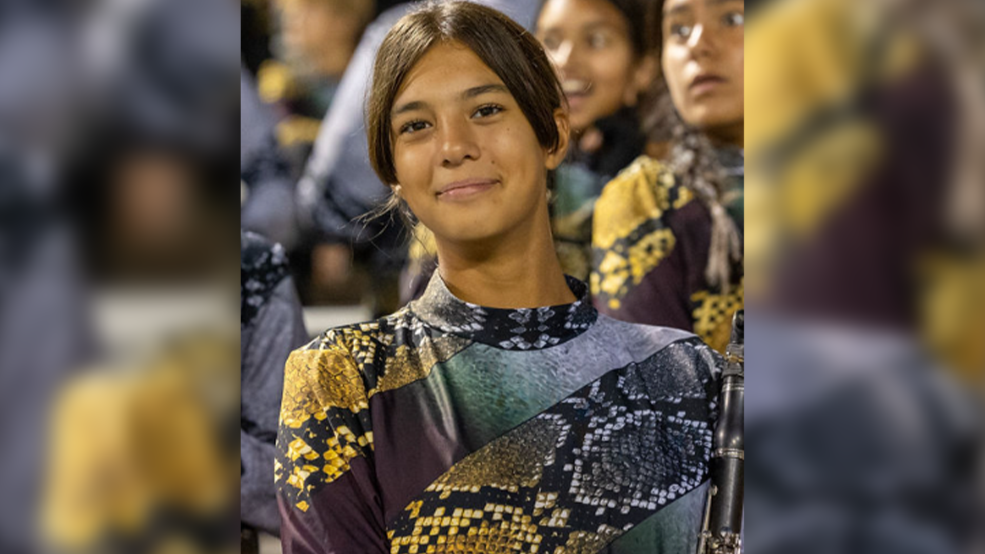 A young woman with dark hair and a patterned dress stands in a crowd of people, her expression thoughtful and engaged.