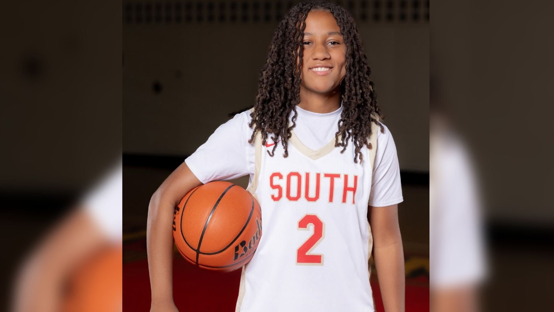 The image shows a person wearing a white basketball jersey with the word "SOUTH" and the number "2" printed on it, holding a basketball. The person has long dreadlocks and is standing in what appears to be a dimly lit indoor setting.