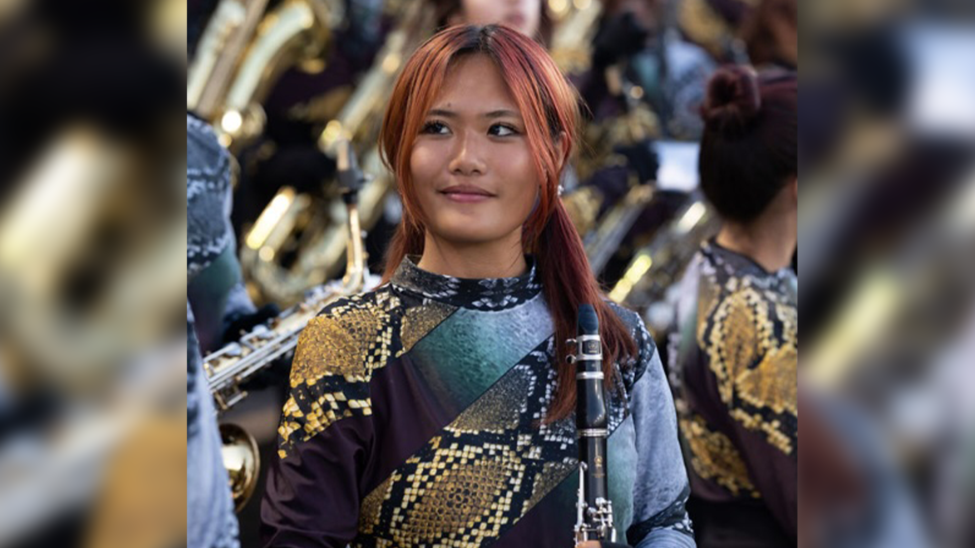 A young woman with vibrant red hair and a patterned jacket stands in a crowded, dimly lit setting.