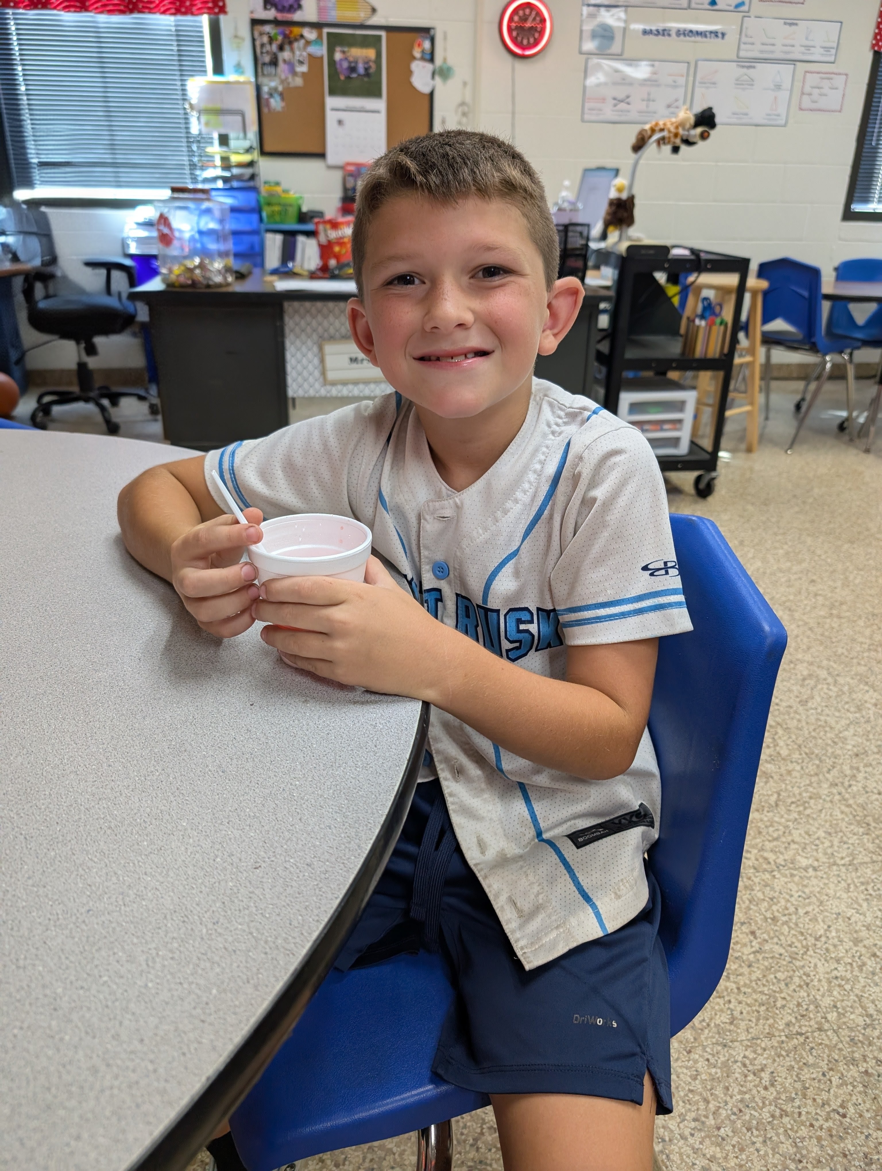 A young boy in a school uniform is sitting at a desk, holding a cup and smiling at the camera. The classroom environment with various educational materials and posters can be seen in the background.