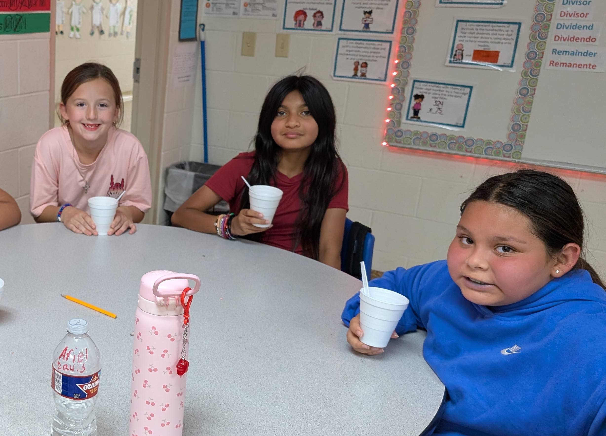Three young students, two girls and one boy, are sitting at a table in a classroom setting, each holding a cup or bottle.