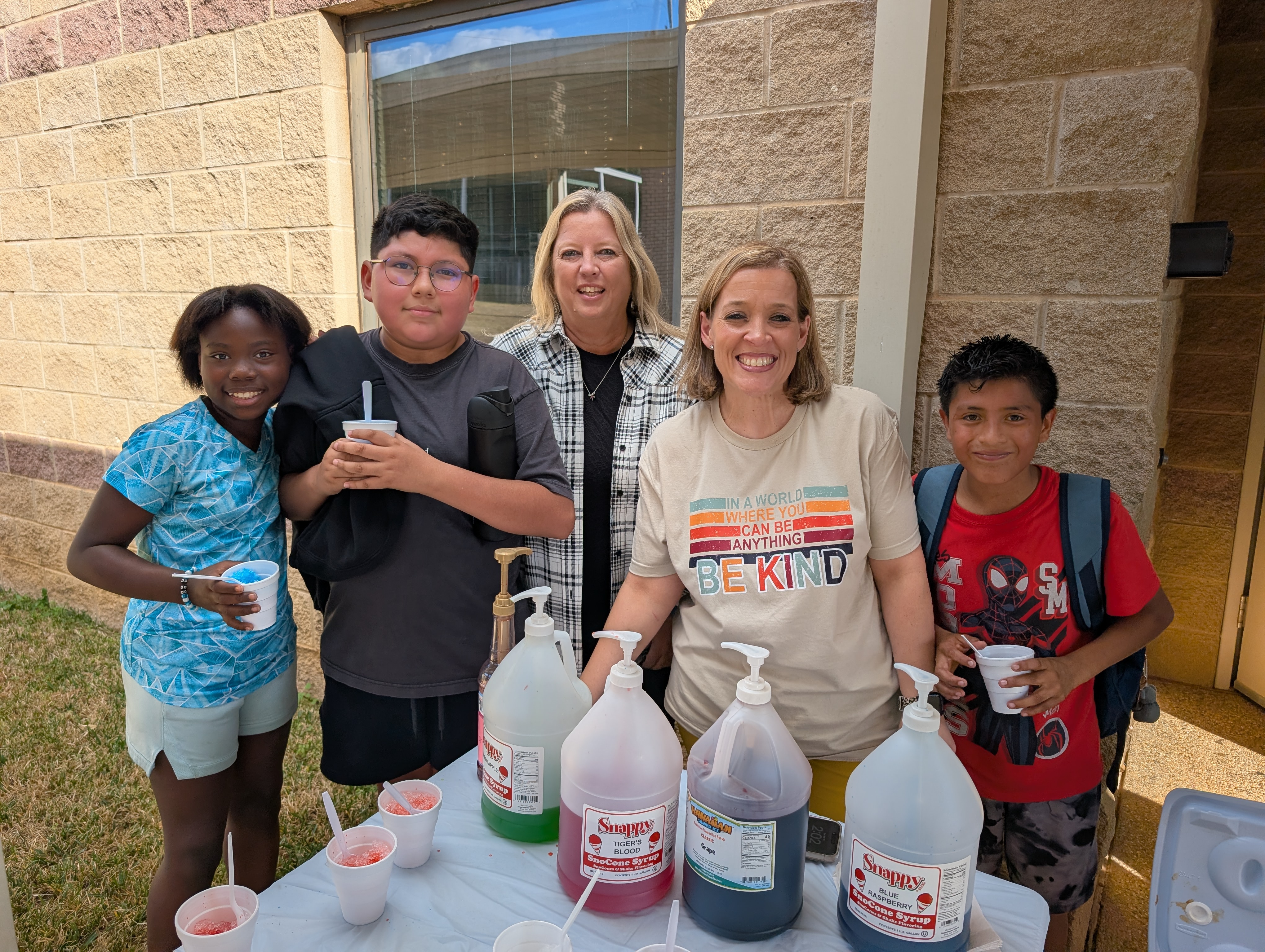 A group of people, including two adults and three children, are standing in front of a building holding various items, including cleaning supplies and drinks.