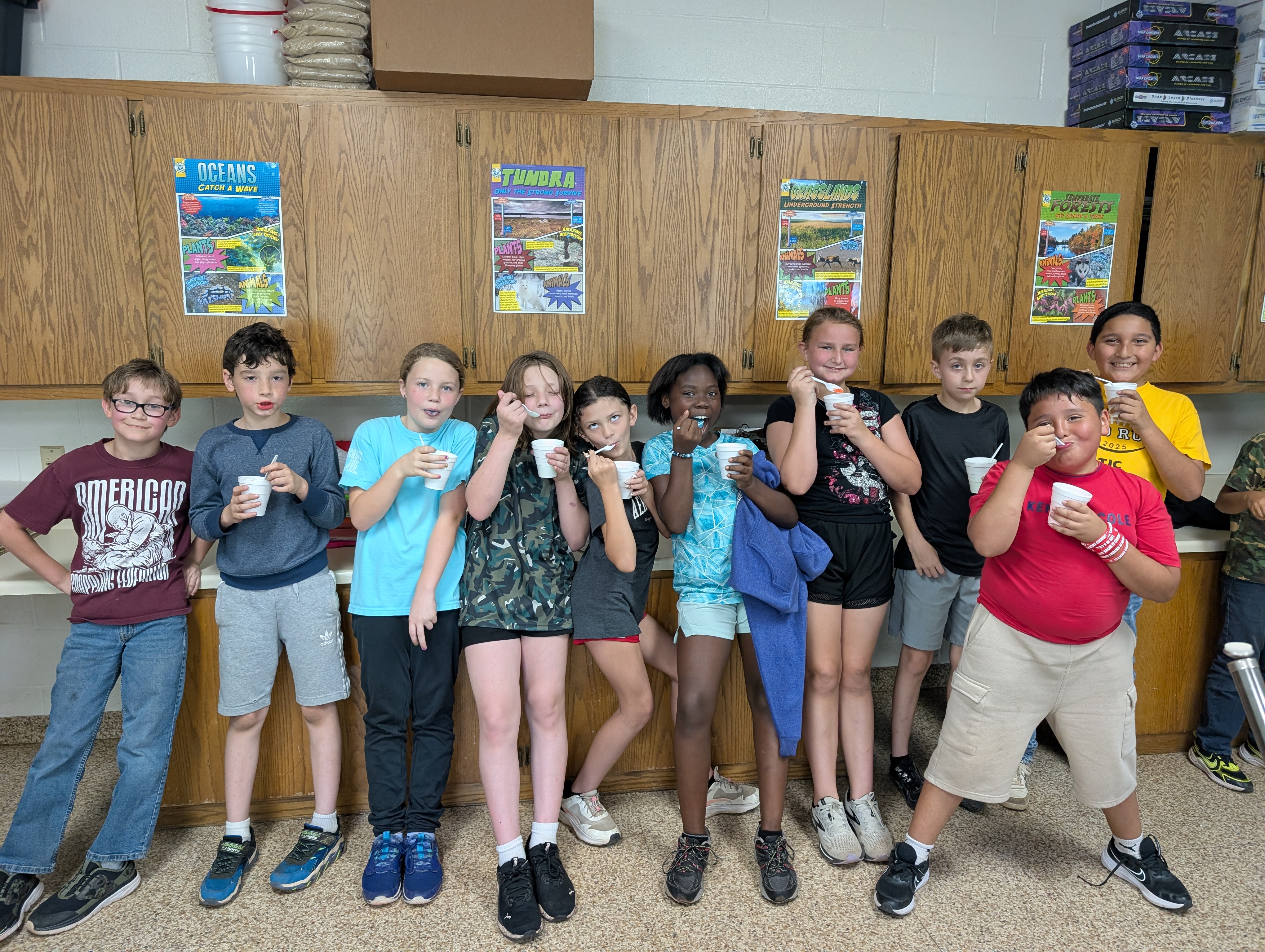 A group of young people, both boys and girls, standing together in what appears to be a classroom or community center setting, with various posters and shelves visible in the background.