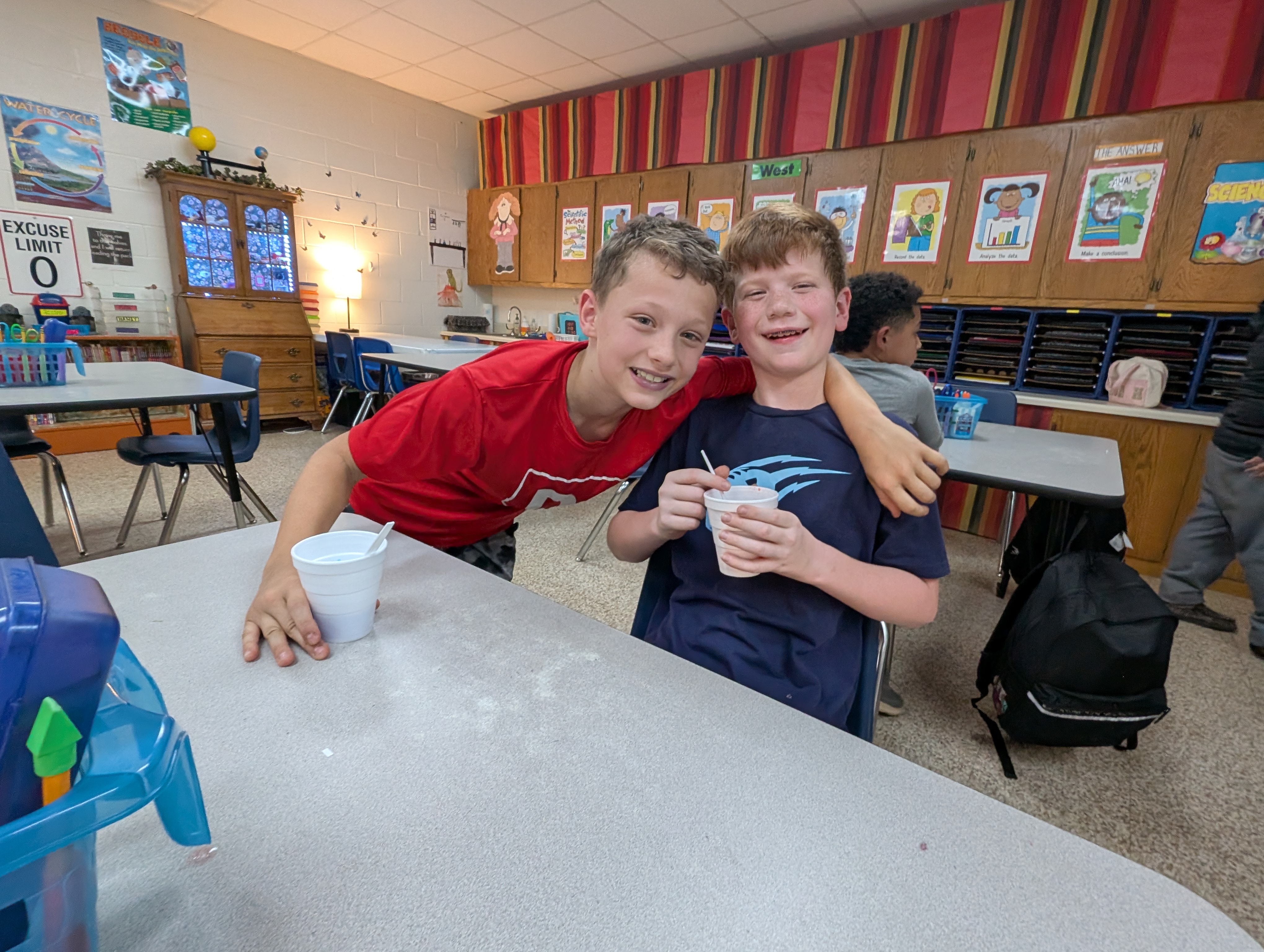Two young boys, one wearing a red shirt and the other a blue shirt, are sitting together at a table in what appears to be a classroom setting. The background includes colorful wall decorations and shelves, suggesting an educational environment.
