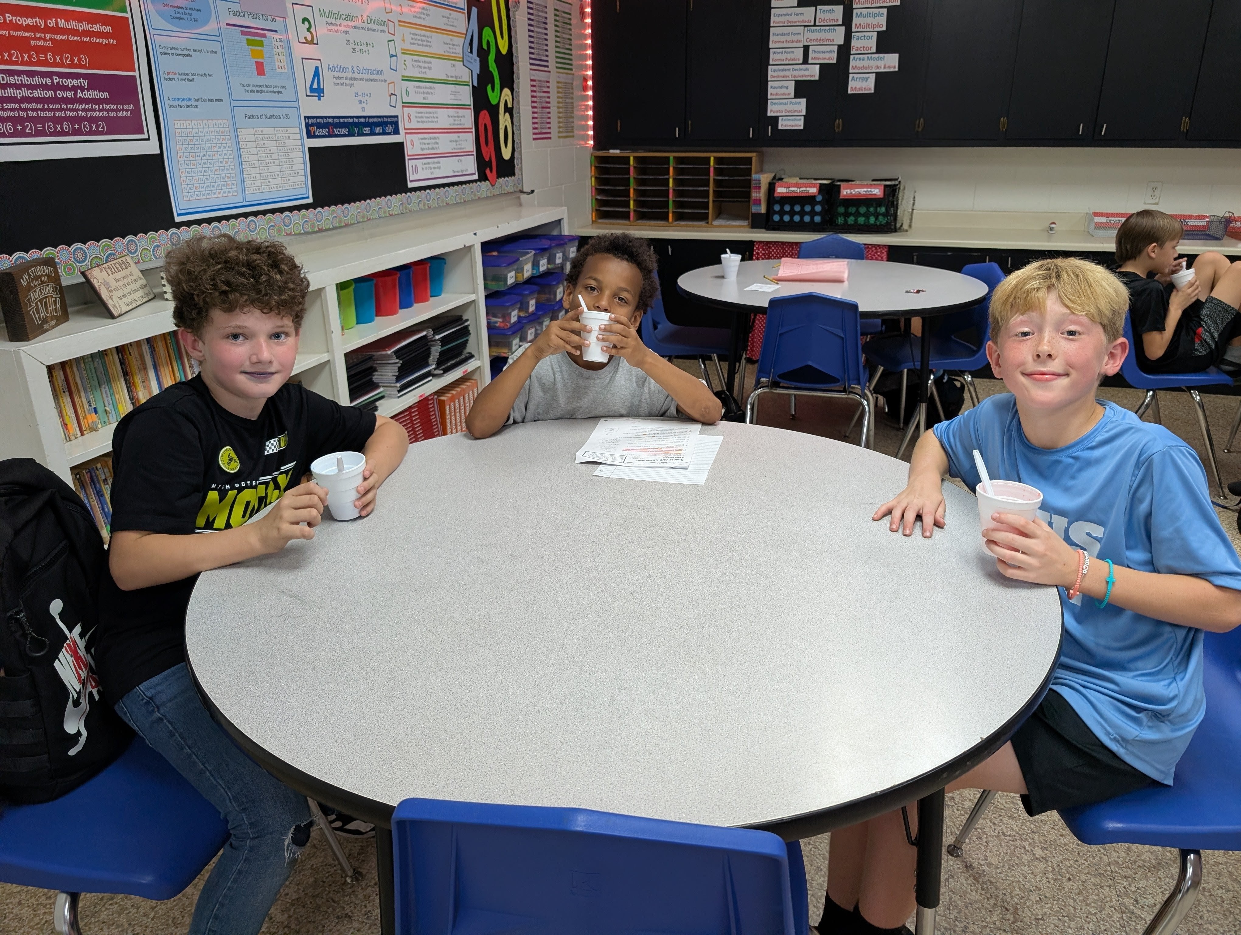 Three young students are sitting at a table in a classroom, each holding a drink and appearing to be engaged in conversation or an activity.