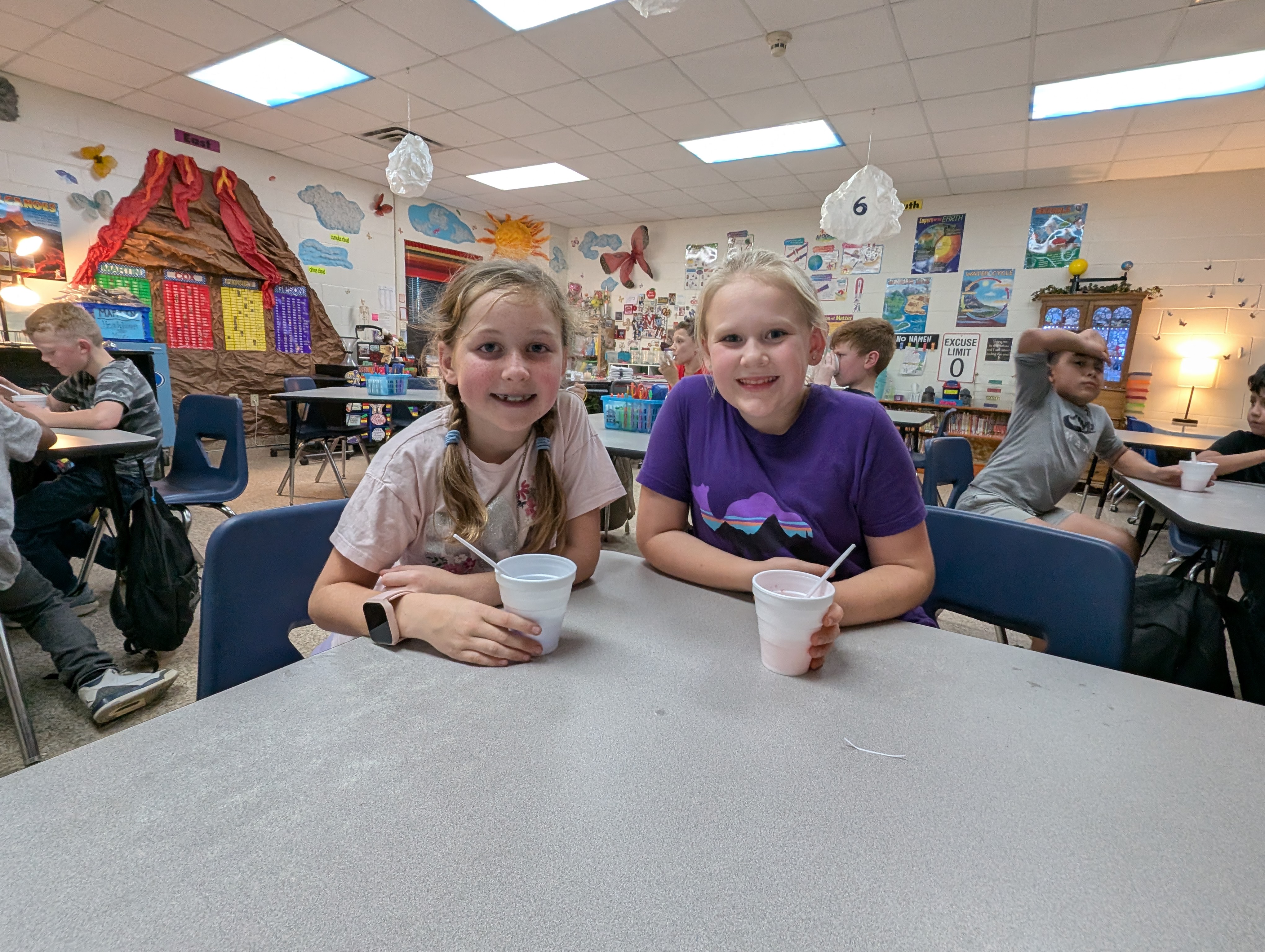 Two young girls, one wearing a pink shirt and the other a purple shirt, are sitting at a table in a colorful and lively classroom environment filled with various decorations and artwork on the walls.
