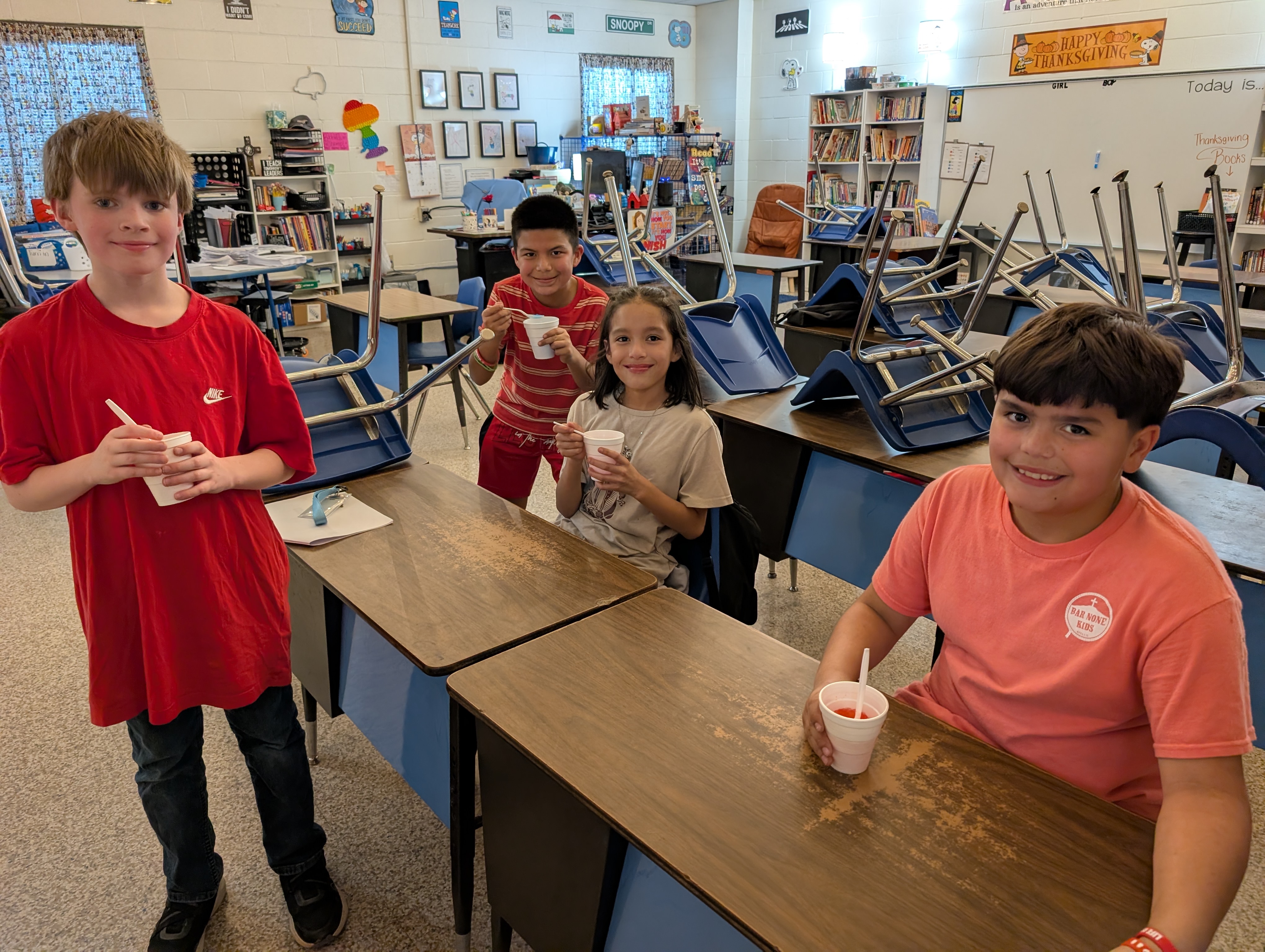 The image shows a group of young students in a classroom setting, with various educational materials and furniture visible in the background.