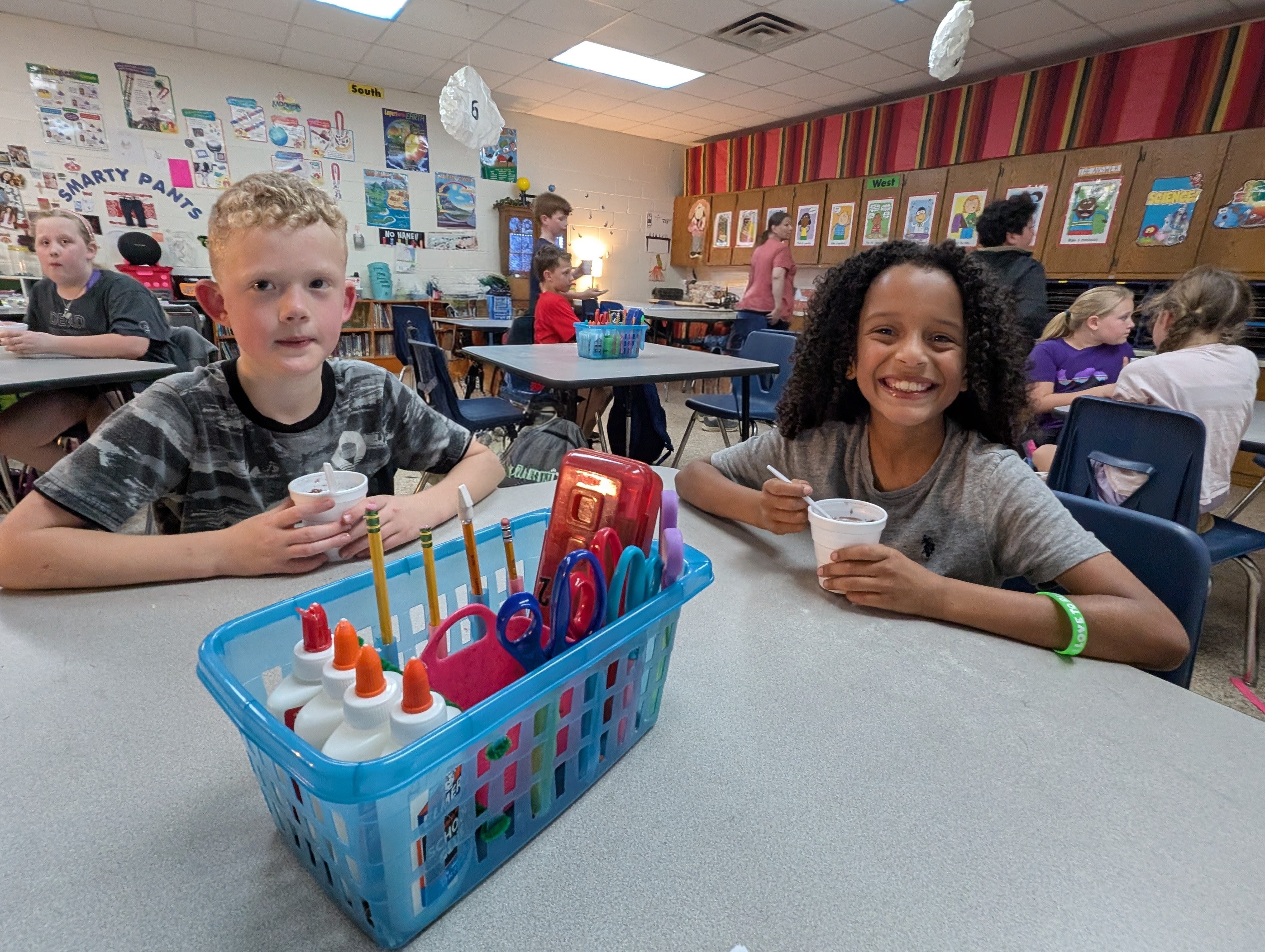 The image shows two students, a boy and a girl, sitting at a table in a classroom setting. The boy is holding a small object, and the girl is smiling. The classroom walls are decorated with various posters and artwork, creating a vibrant and engaging learning environment.