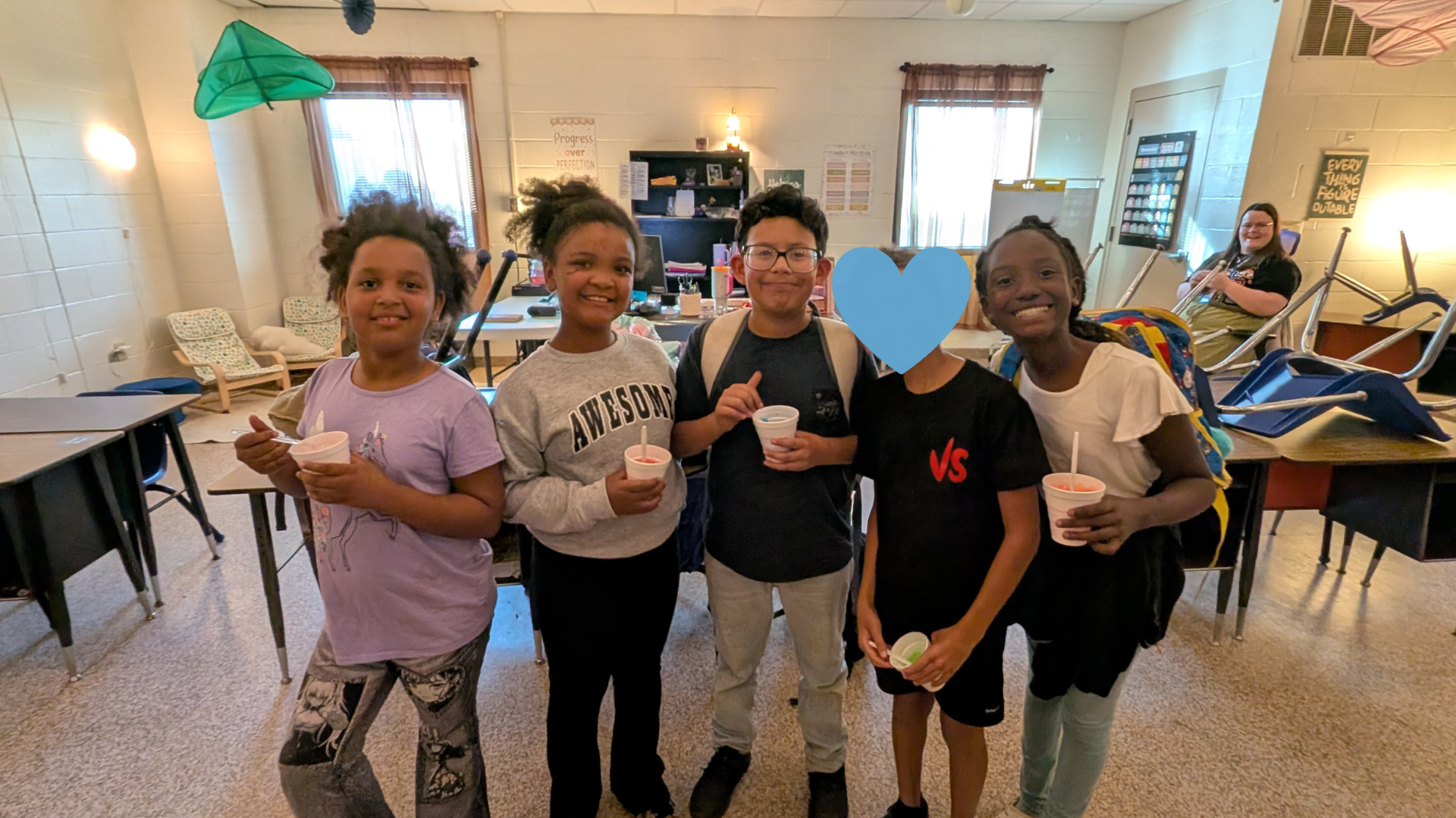 The image shows a group of four young people, likely students, standing together in what appears to be a classroom or educational setting. They are smiling and holding various items, such as a green triangle and a blue shape, suggesting they may be engaged in some kind of activity or project.