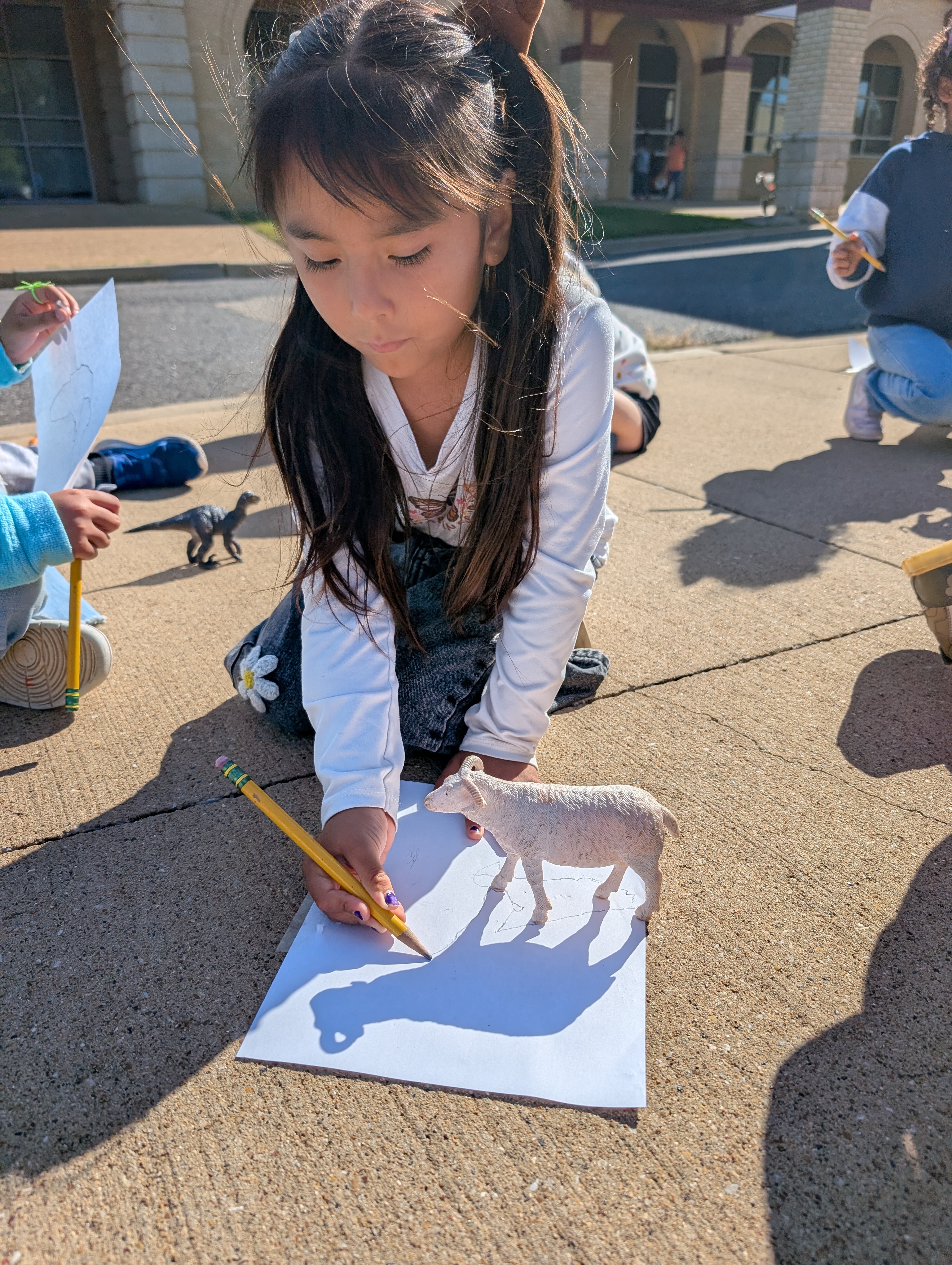 A young girl is intently drawing on a large piece of paper on the ground, surrounded by various toys and objects in an outdoor setting.