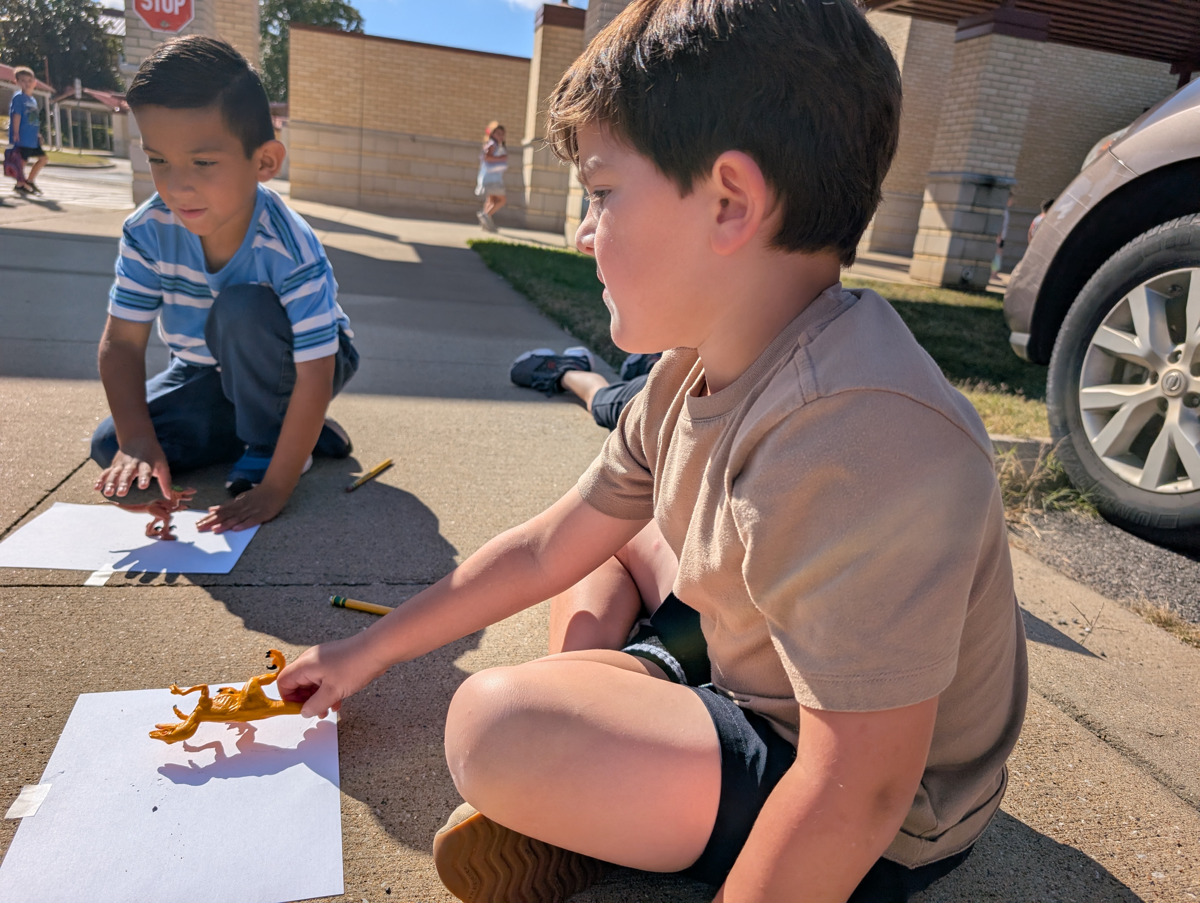 Two young boys are sitting on the sidewalk, engaged in an activity that appears to involve drawing or painting. In the background, there is a building and a parked vehicle.