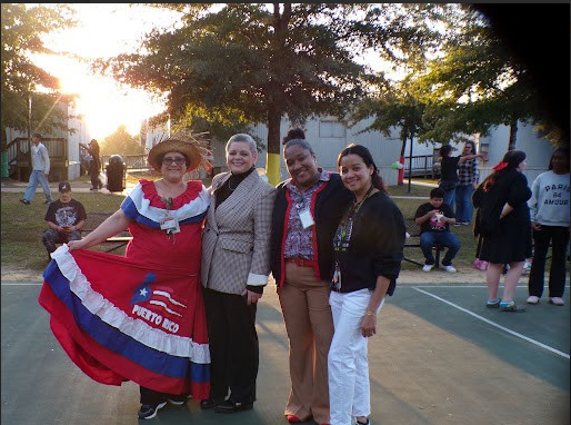 A group of four women, dressed in colorful and festive attire, stand together in a park-like setting with trees and other people visible in the background.