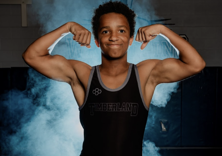 A young man with curly hair is flexing his muscular arms in front of a blue smoky background, wearing a black tank top with the word "Neverland" printed on it.