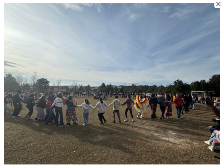 A large group of people, some holding hands, standing in a field with trees and a cloudy sky in the background.
