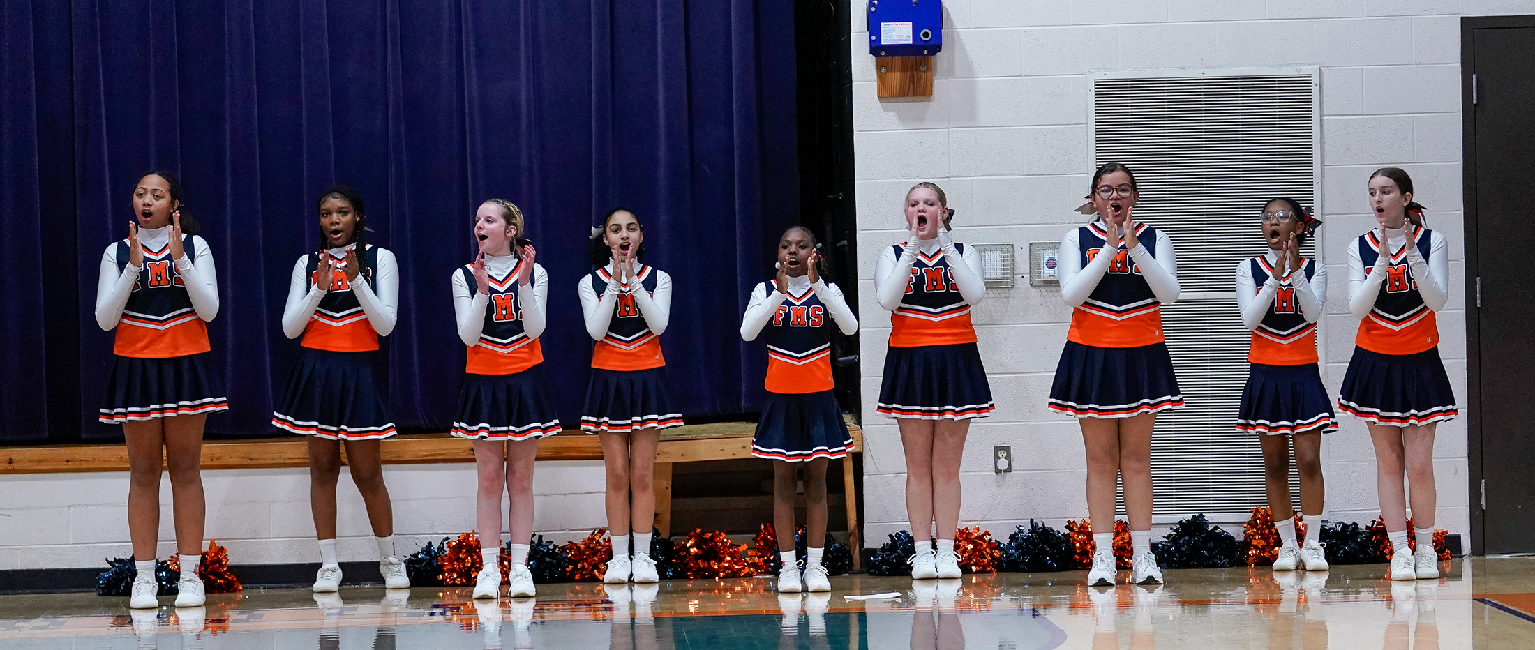 cheerleaders in blue and orange uniforms clap in a gym
