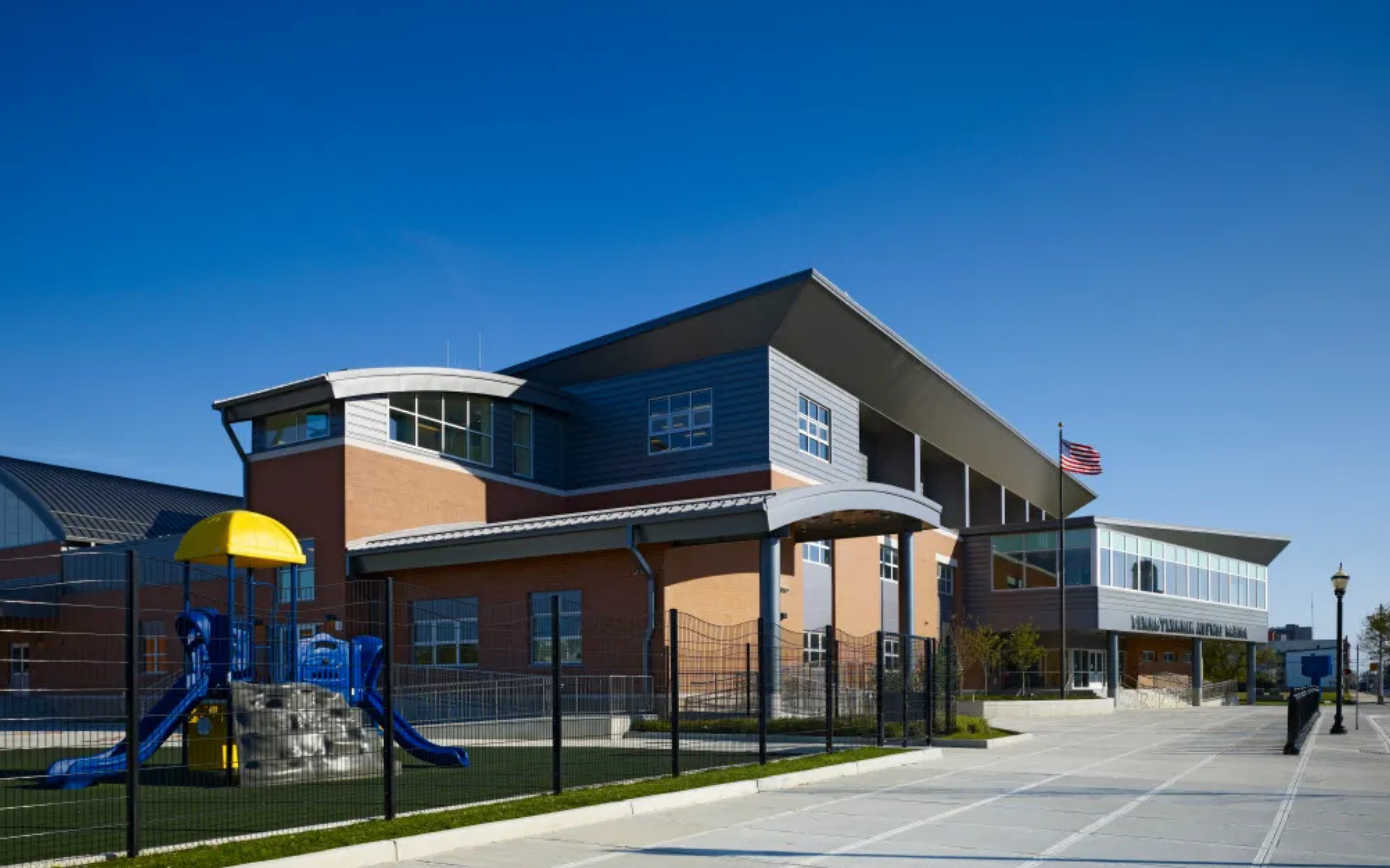 A colorful children's playground sits behind a black fence in the foreground, with a modern multi-story school building and a clear blue sky in the background.