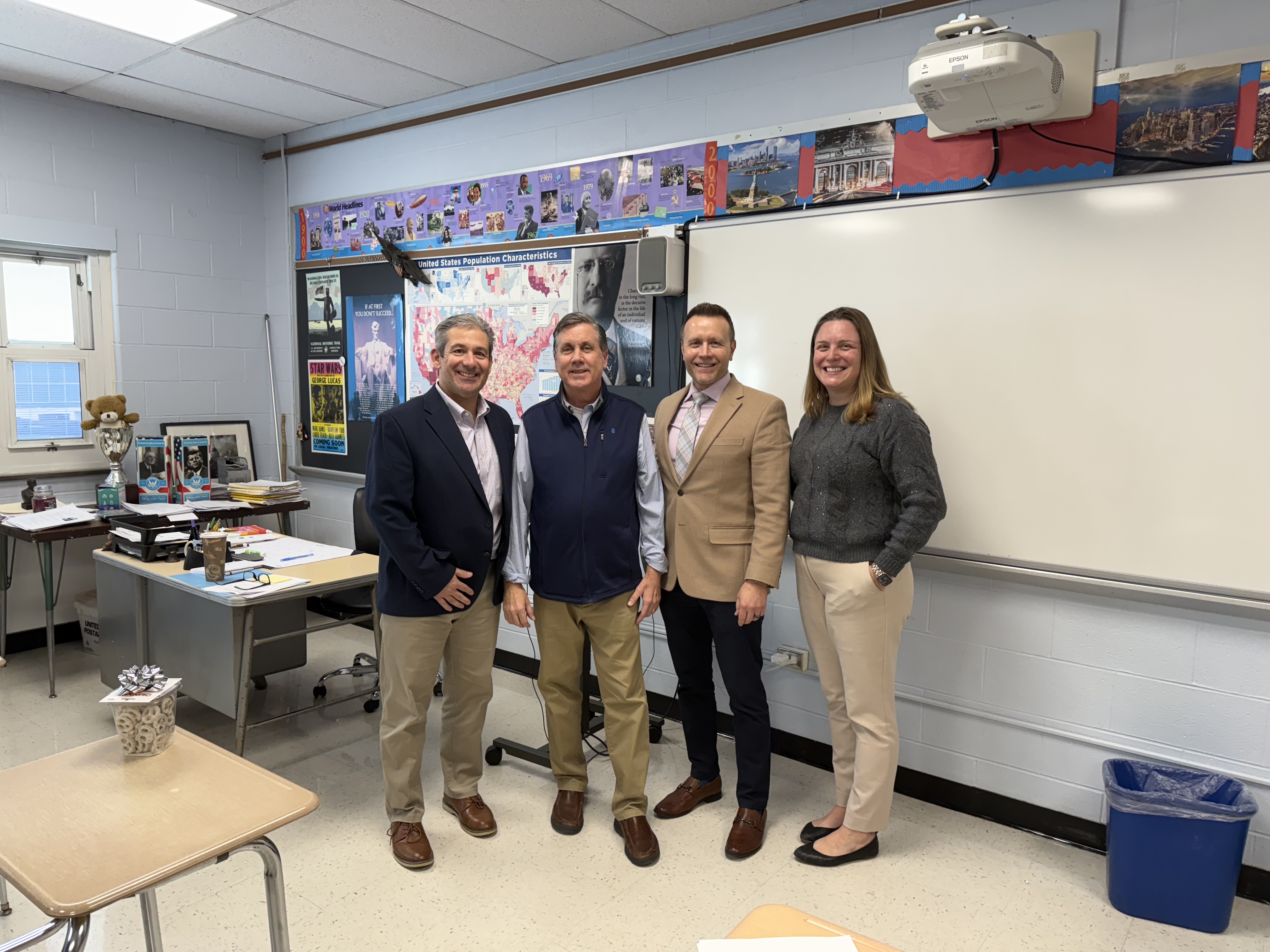Four adults stand in a classroom with a whiteboard and desks in the background, and a table with a gift in the foreground.