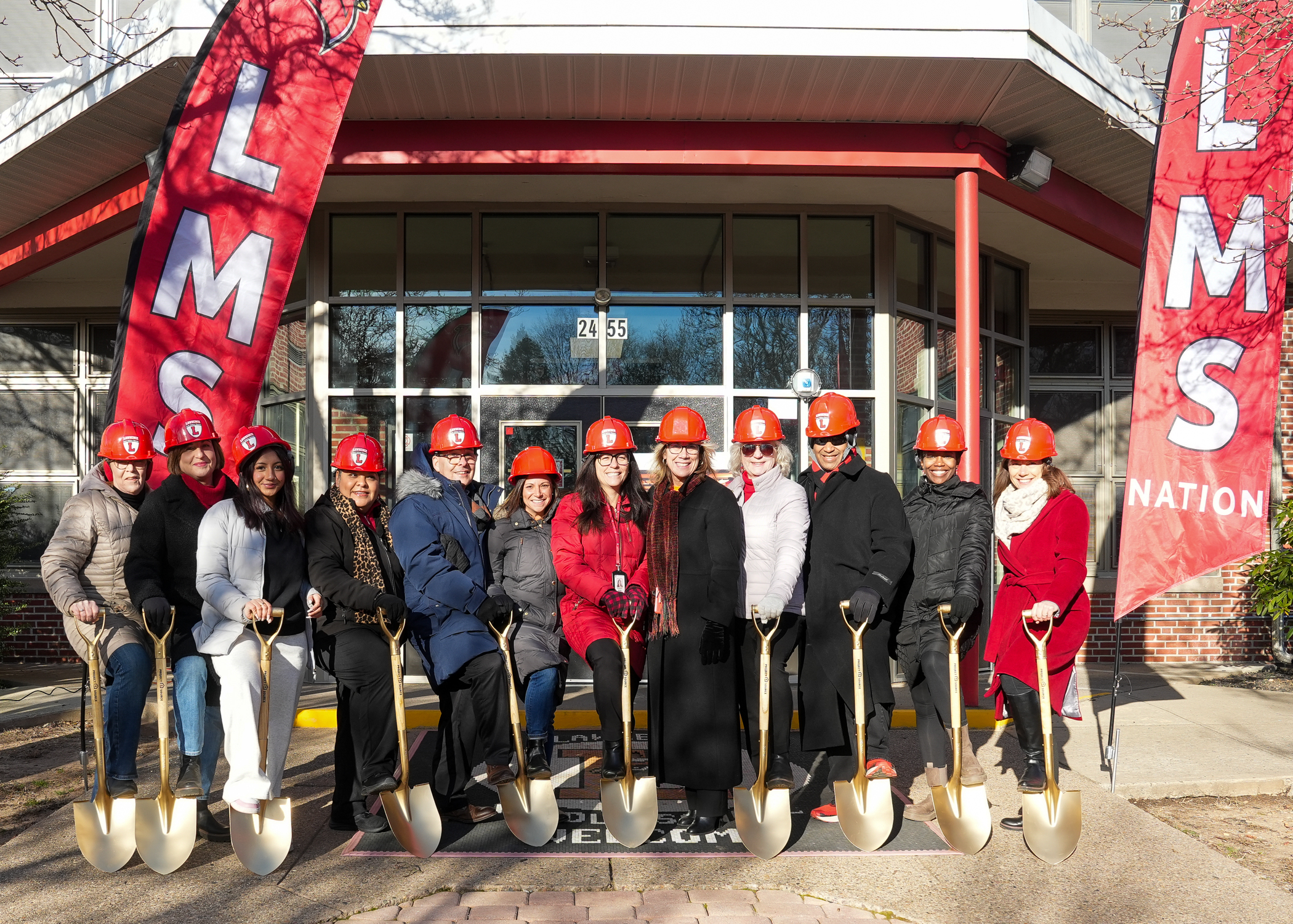 A group of people in hard hats and construction gear stand in front of a building with "LMNS Nation" signage, holding shovels and posing for a photograph.