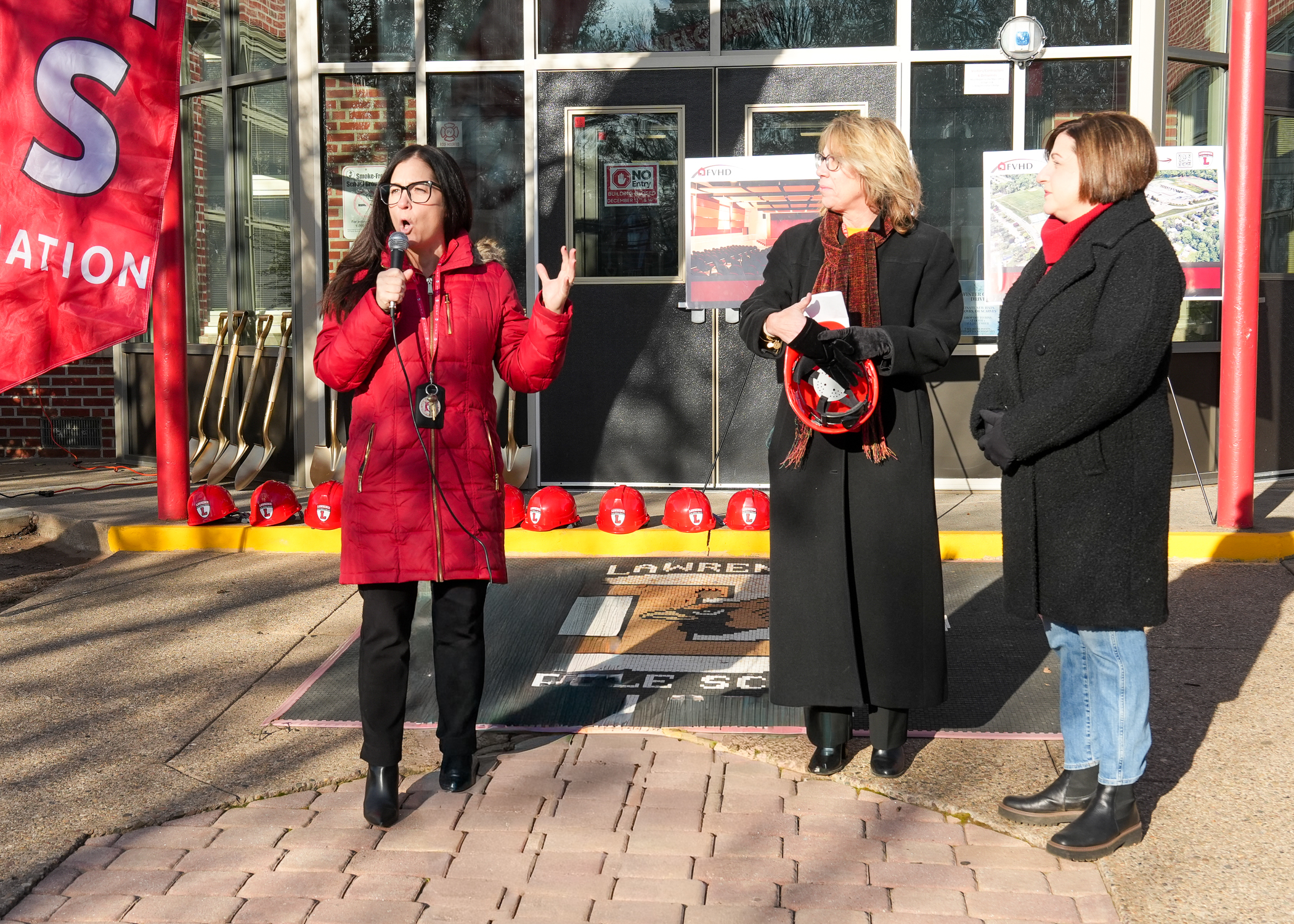 Three women in winter coats standing in front of a storefront with a large "S" sign, against a backdrop of brick buildings and a snowy street.
