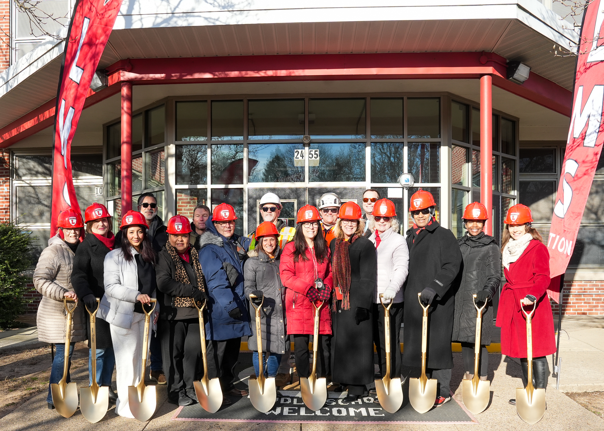 A group of people in hard hats and construction attire stand in front of a building with a large "Welcome" sign, suggesting a groundbreaking or construction event.