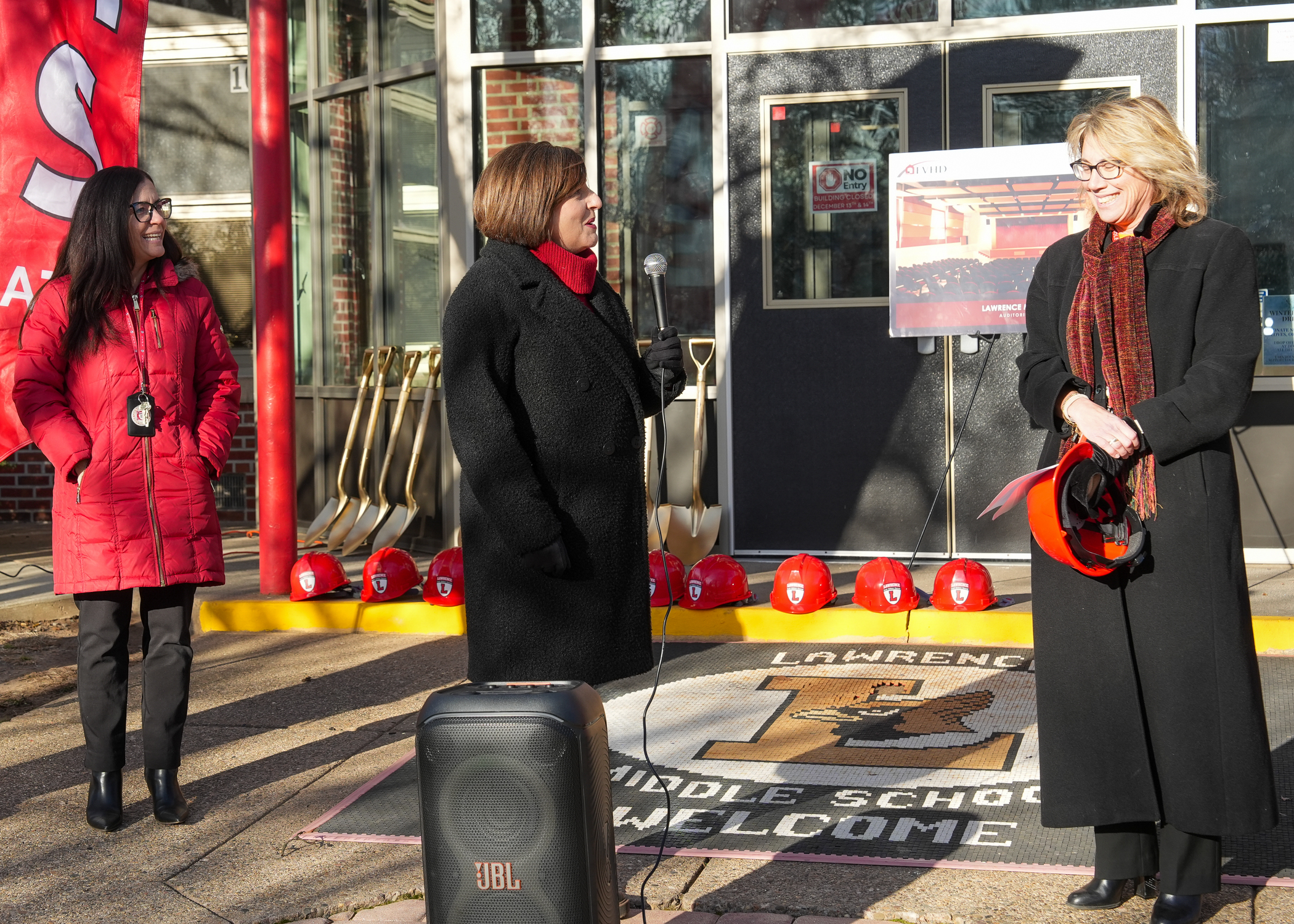 Three individuals, two women and one man, are standing in front of a building with a "Lawrence Middle School Welcome" sign displayed in the foreground.